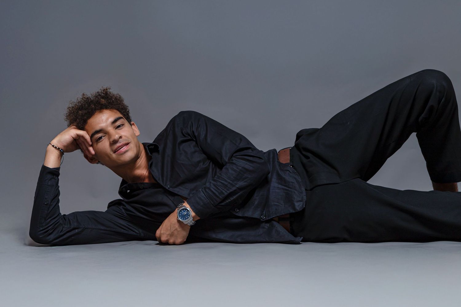 Young man in all-black outfit lying on floor, wearing a blue watch, posing against a gray studio background.
