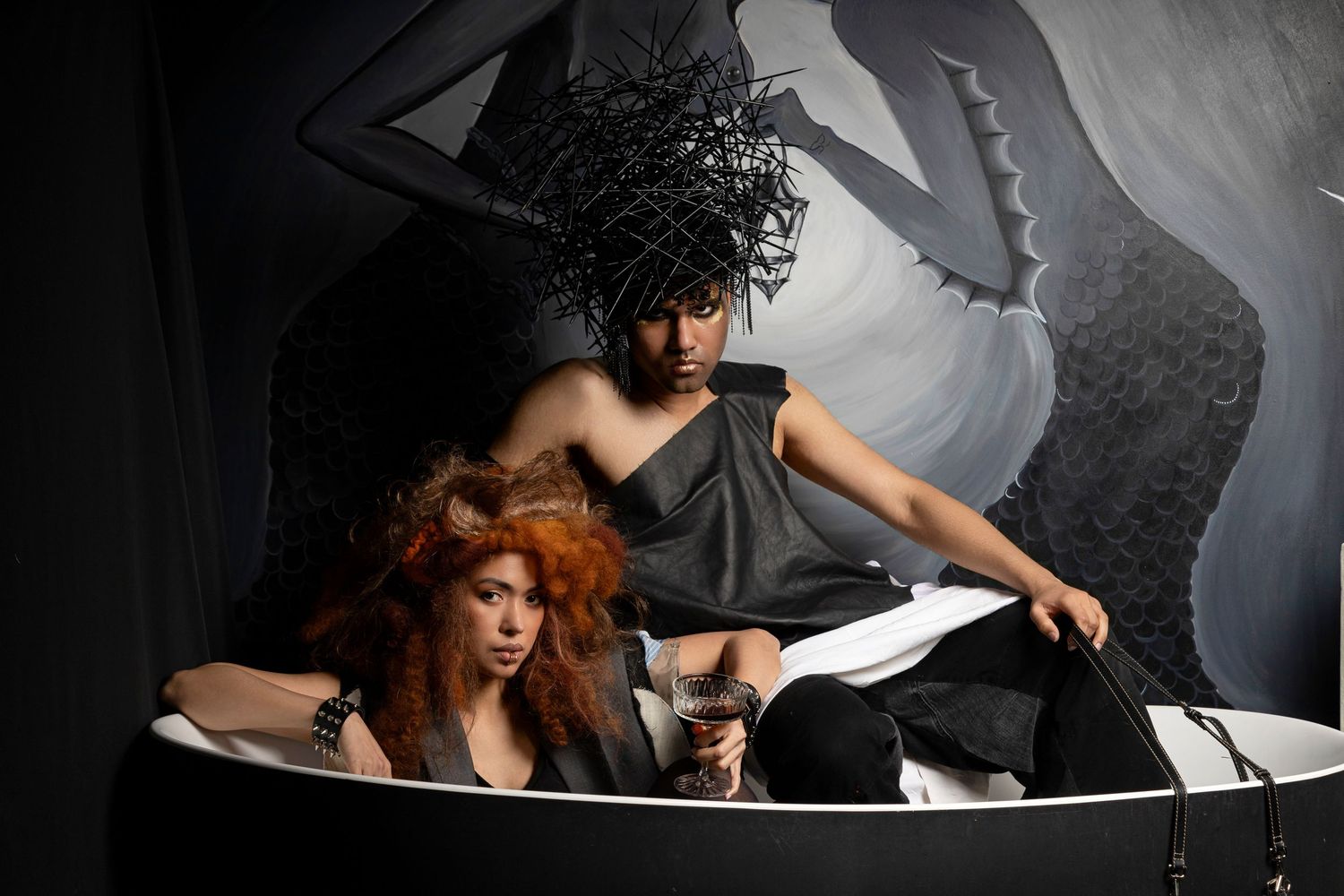 Two models in dramatic black fashion pose in a white bathtub against a dark artistic backdrop.