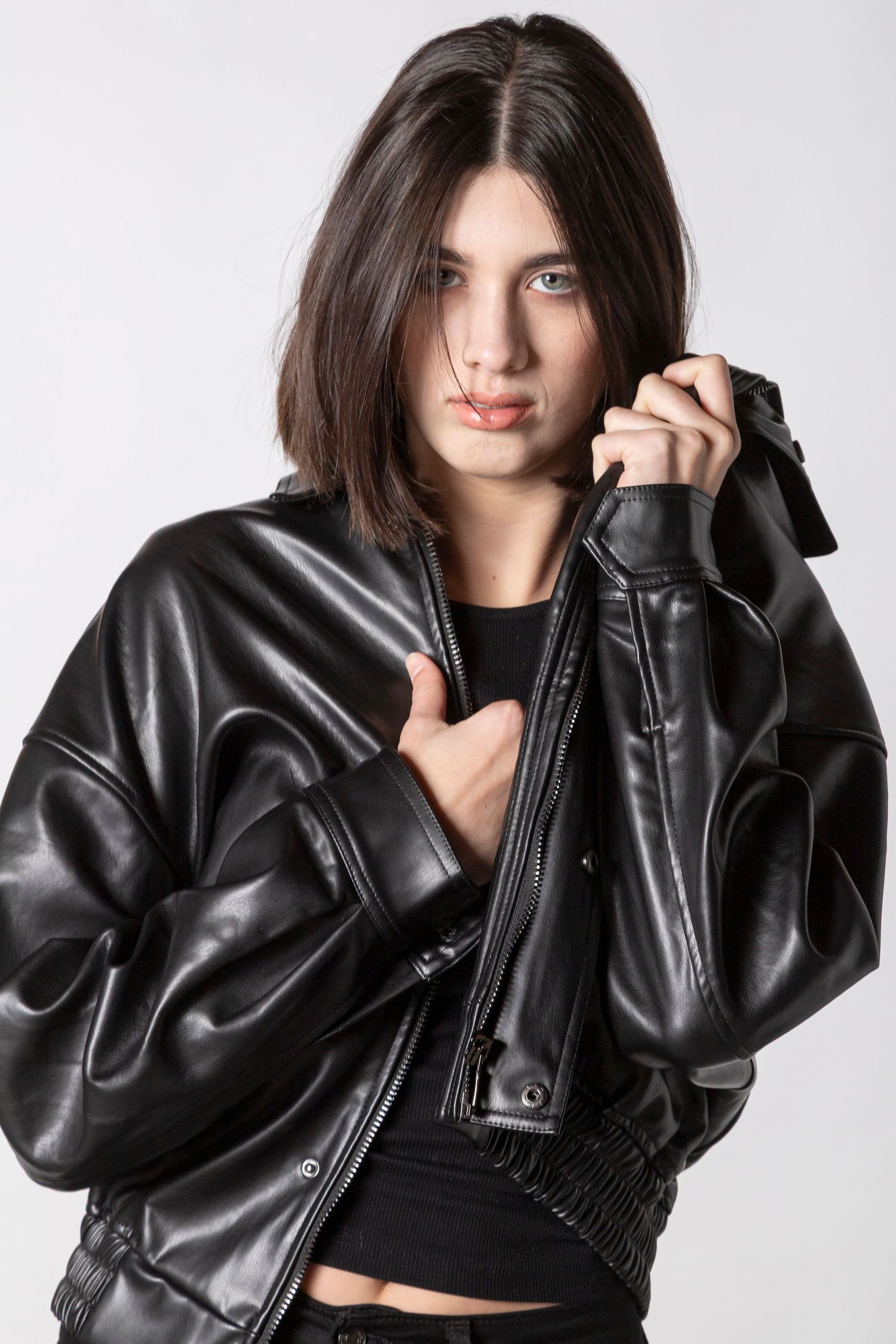Young woman in oversized black leather bomber jacket posing against light gray background with dark hair and blue eyes.