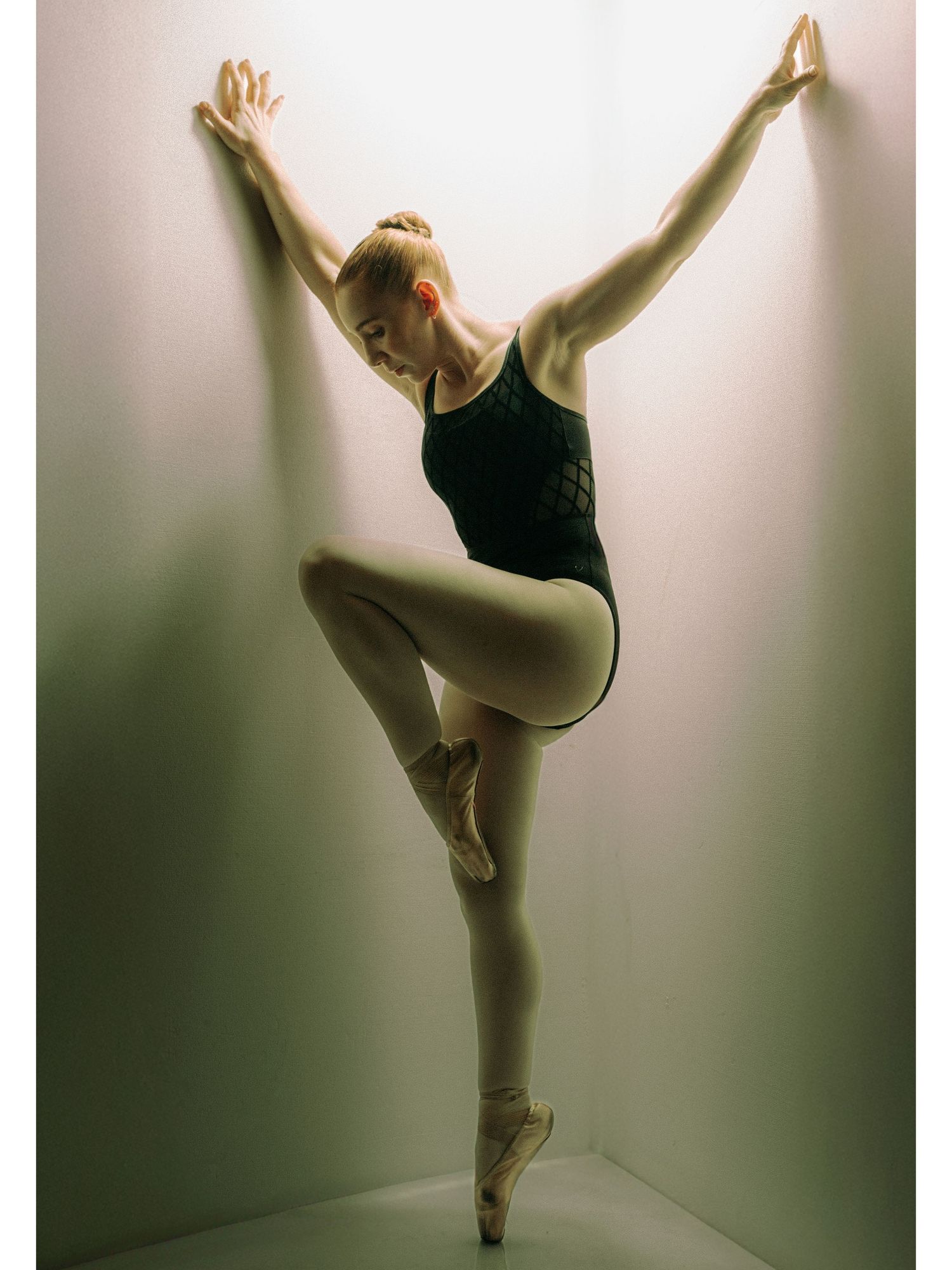 Female ballet dancer in a dark leotard performing an elegant pose on pointe shoes against a wall.