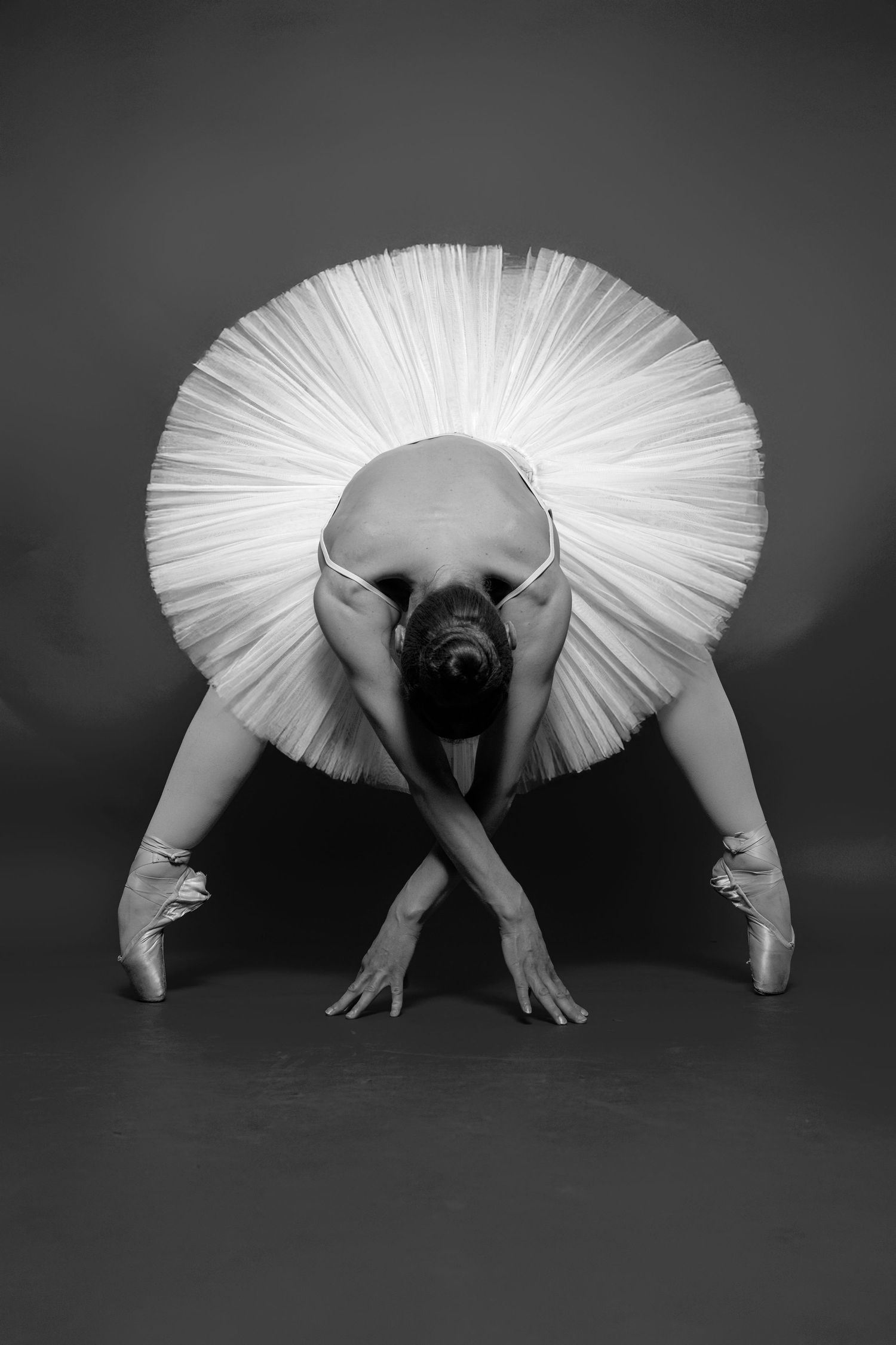 Black and white photo of a ballerina in a white tutu bending forward gracefully in pointe shoes on a dark background.