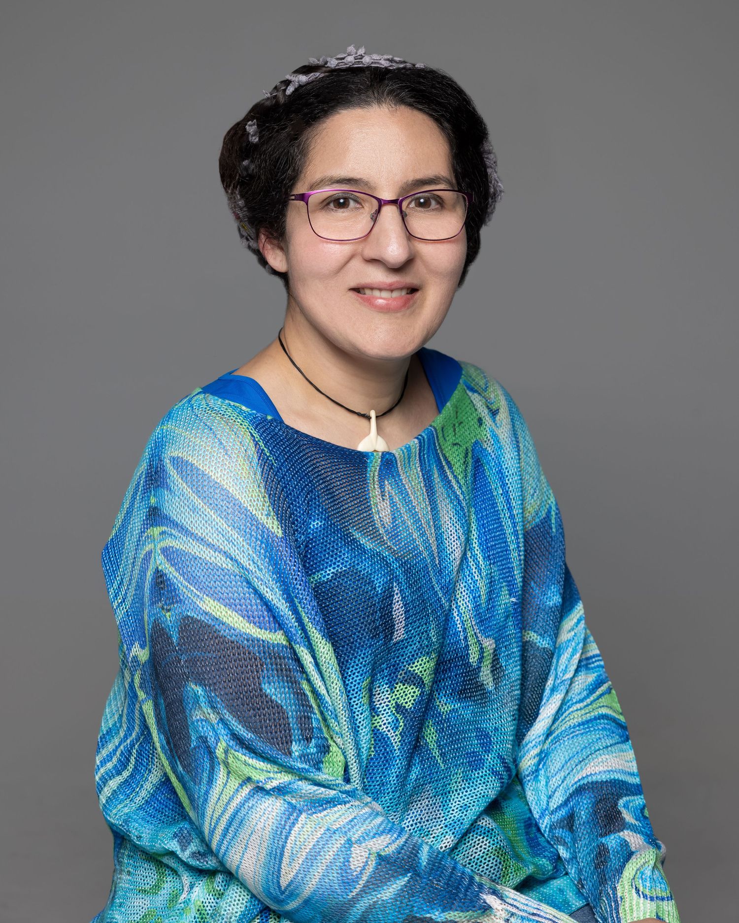 Woman in colorful blue patterned blouse with glasses smiling, wearing a tiara and pendant necklace.