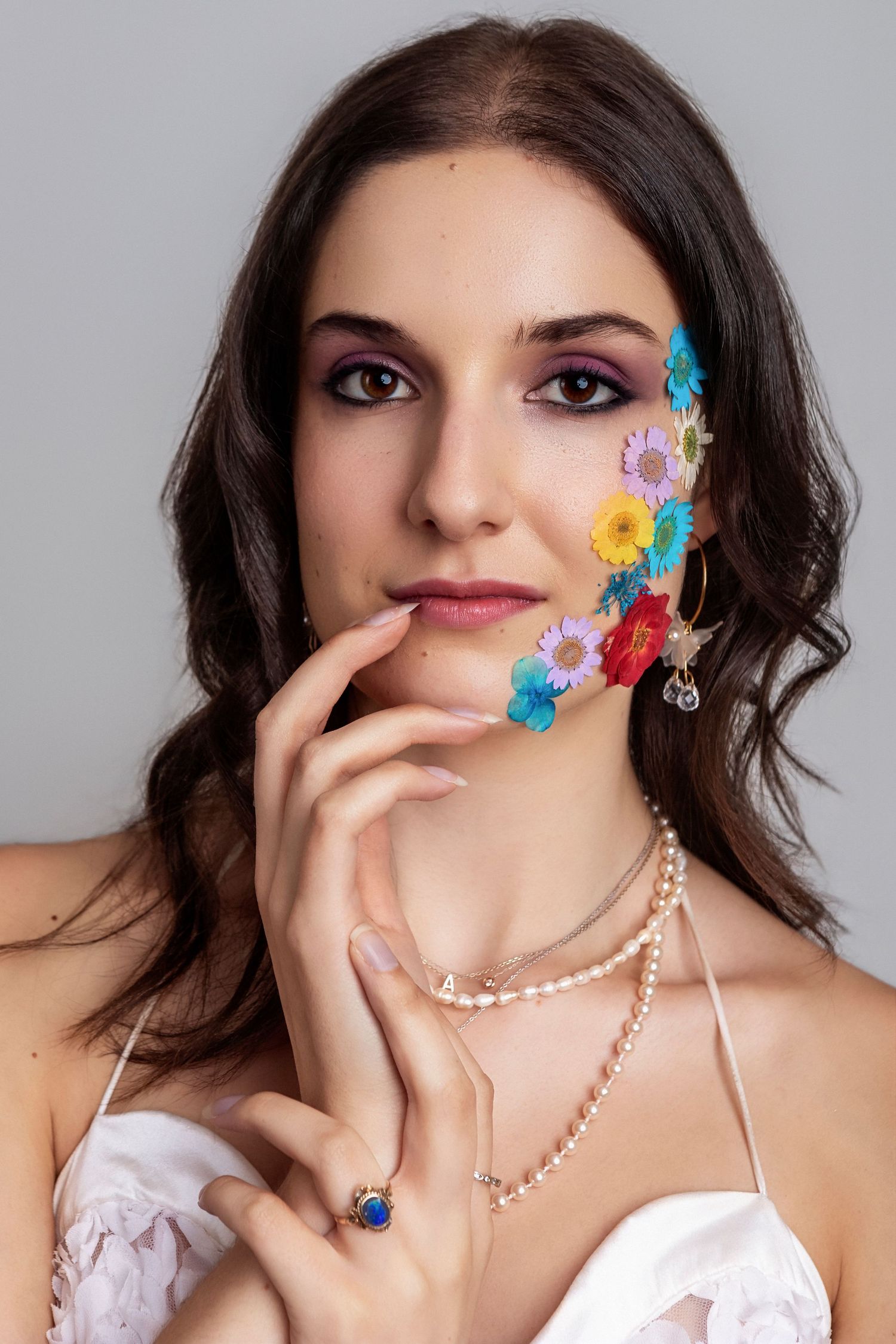Young woman with colorful flowers on cheek, purple eye makeup, pearl necklace, and blue gemstone ring poses elegantly.