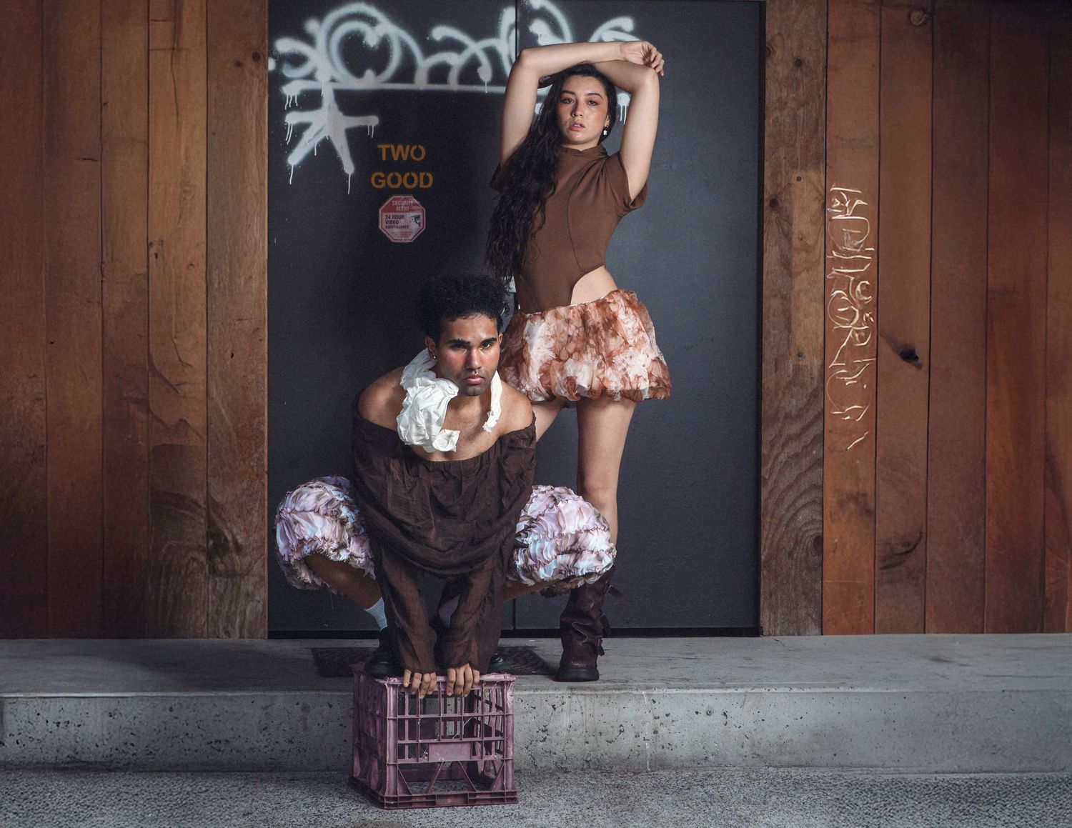 Two dancers pose dramatically in front of a graffiti wall, one crouching on a crate, the other standing above in ruffled skirt.