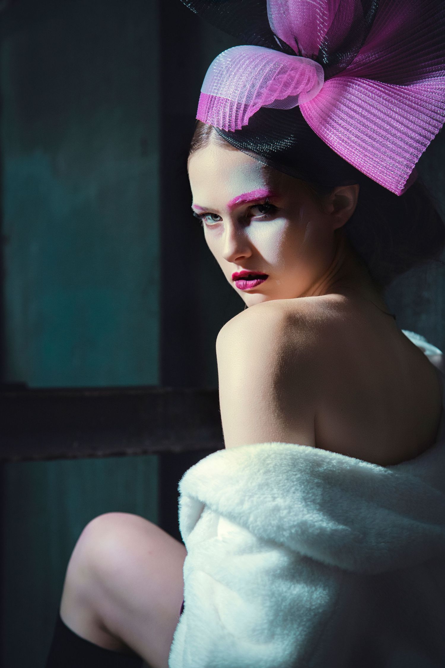 Young woman with dramatic pink makeup and large pink bow fascinator hat, wrapped in white avant-garde outfit, posing in moody dark setting.