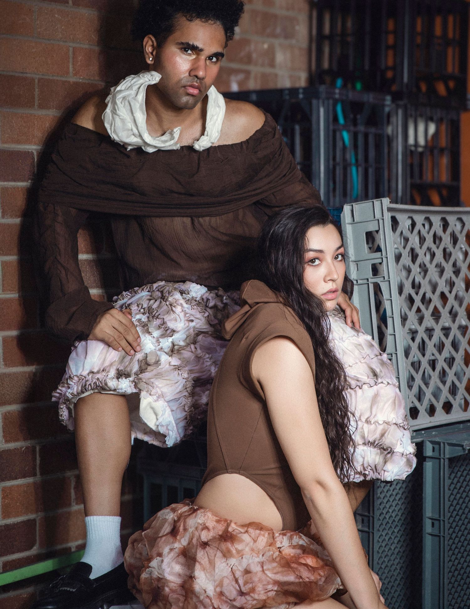 Two models in brown off-shoulder avant-garde tops with ruffled pink floral skirts pose against a brick wall.