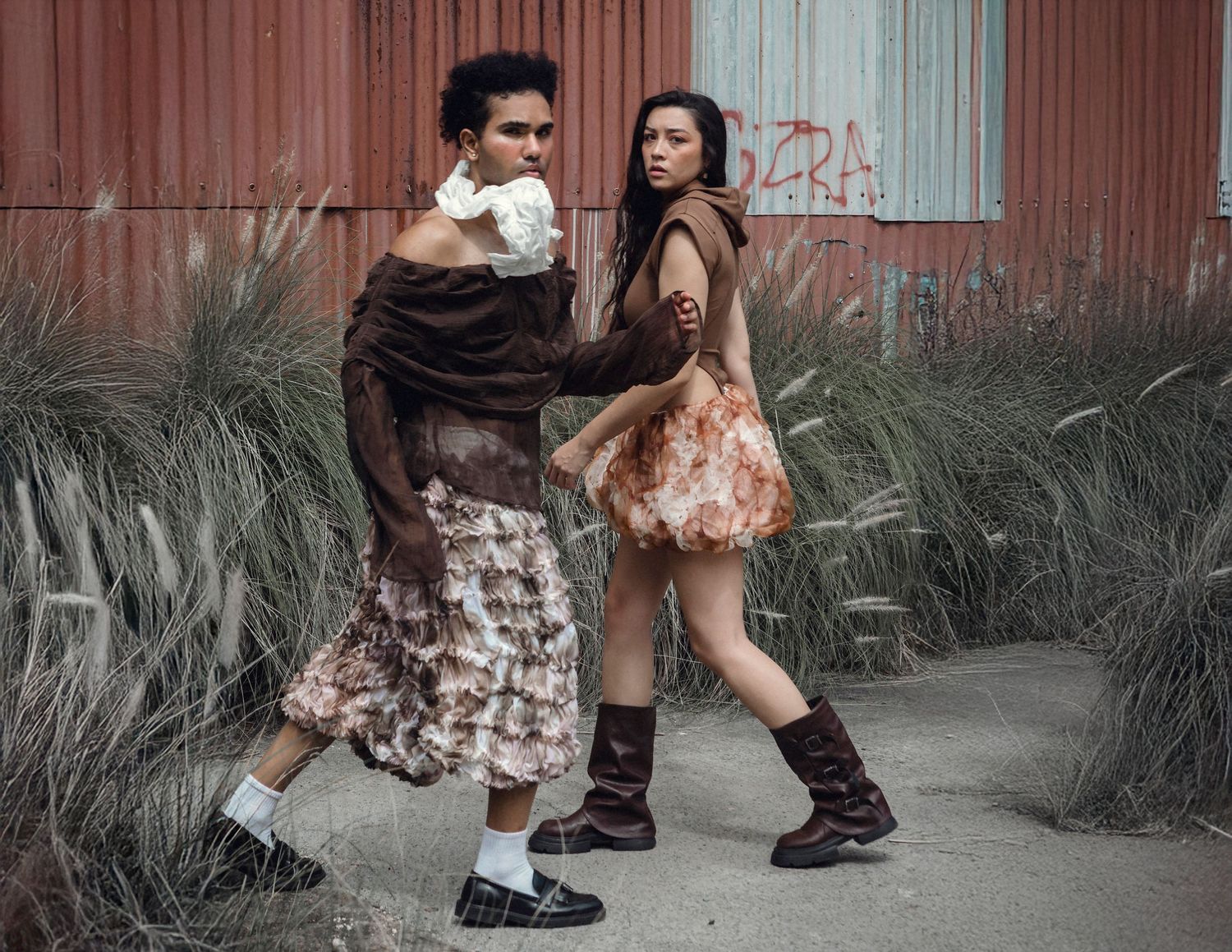 Two models pose in avant-garde brown ruffled outfits and boots against a rustic red building backdrop.
