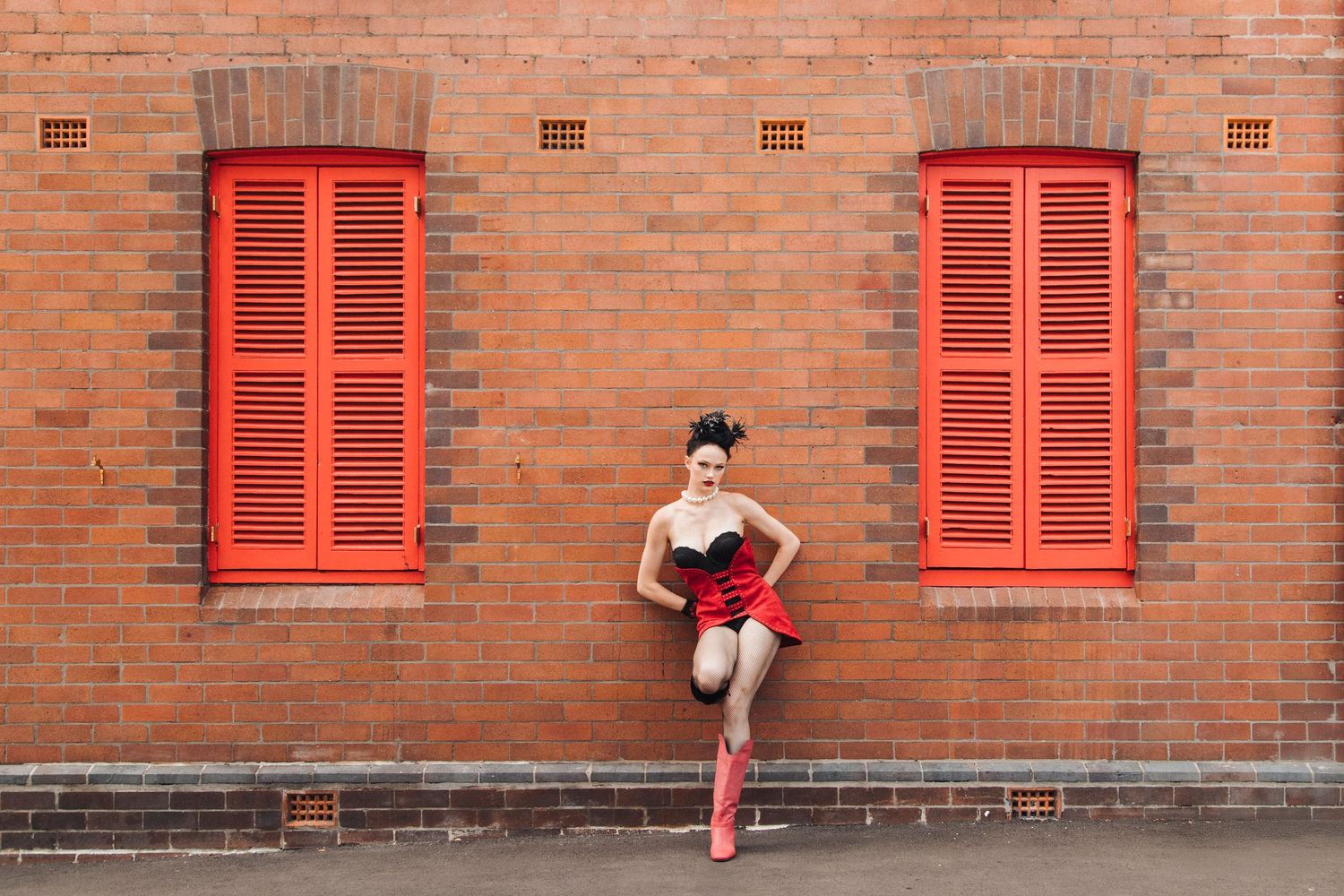 Woman in red skirt and boots posing against a brick wall with red shuttered windows.