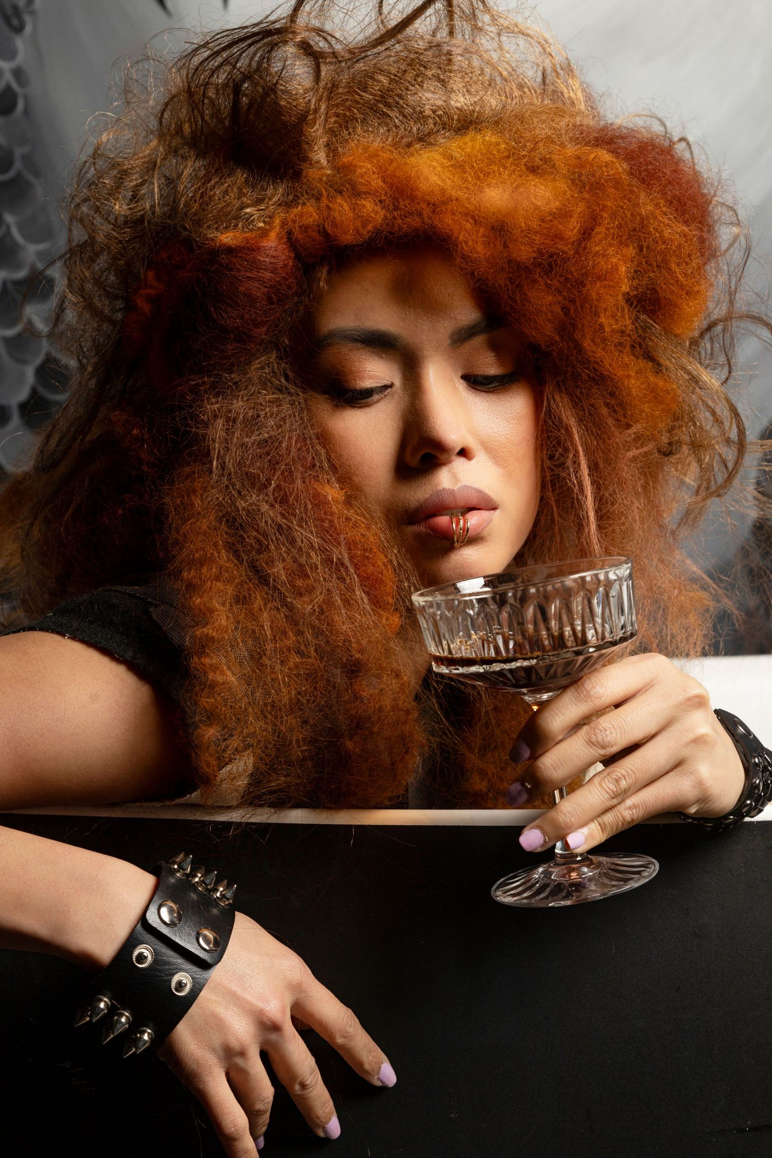 Woman with voluminous red hair holding a crystal glass, wearing a black outfit and studded bracelet.