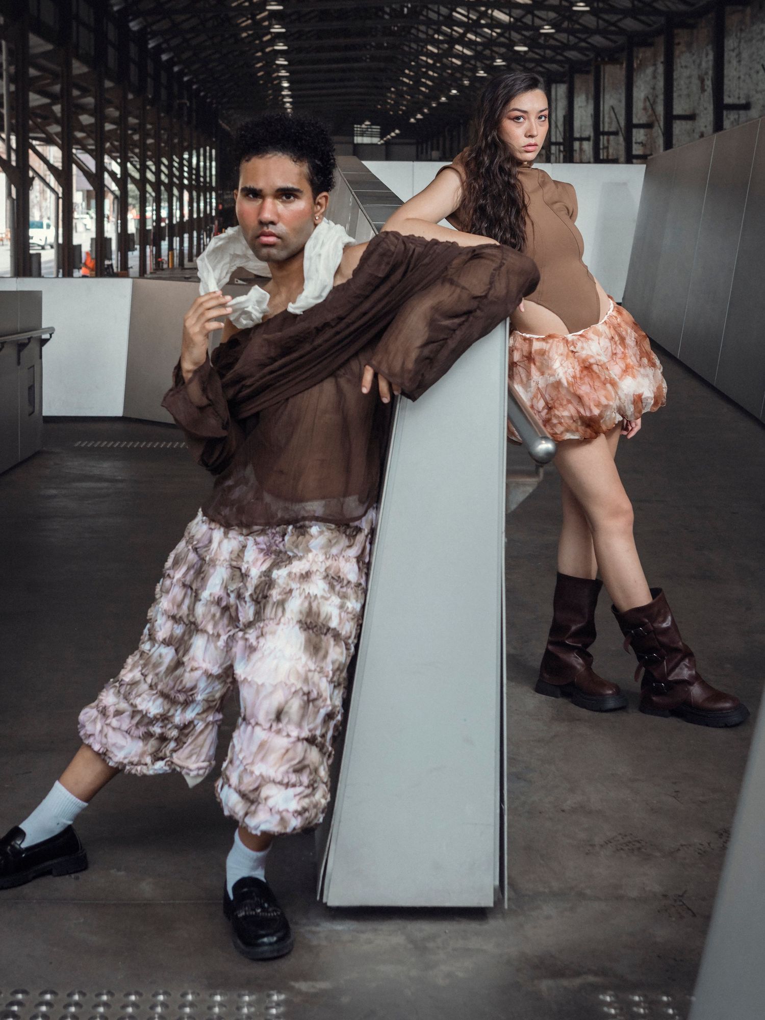 Two fashion models wearing brown and floral outfits posing in a modern industrial corridor.