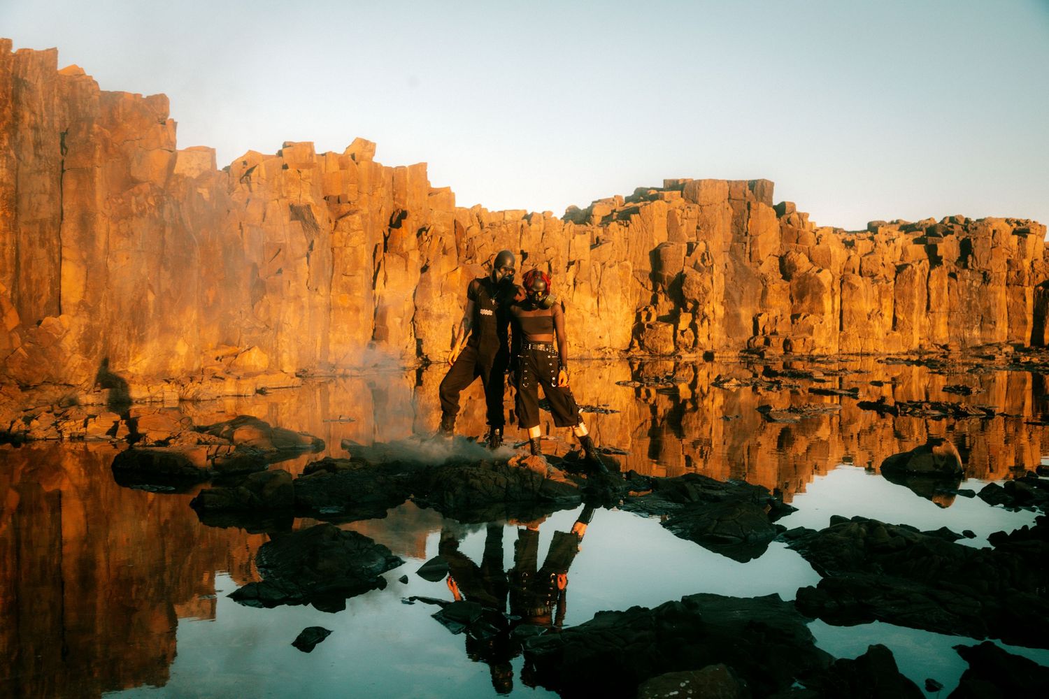 Two people stand on rocks beside a reflective pool at golden hour, surrounded by dramatic sandstone cliffs glowing orange.