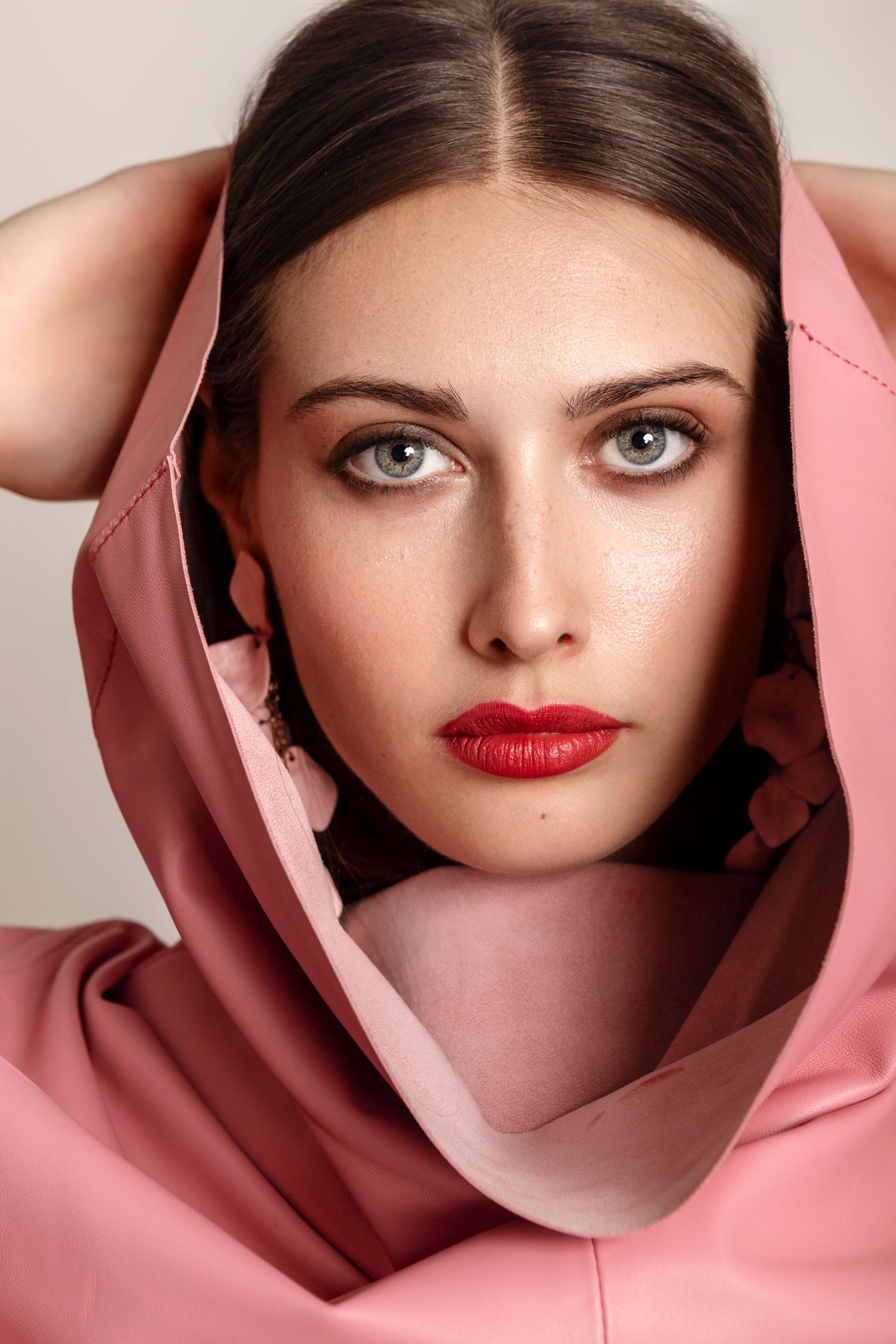 Young woman with green eyes and red lips wearing a pink hooded garment, posing elegantly for a fashion portrait.