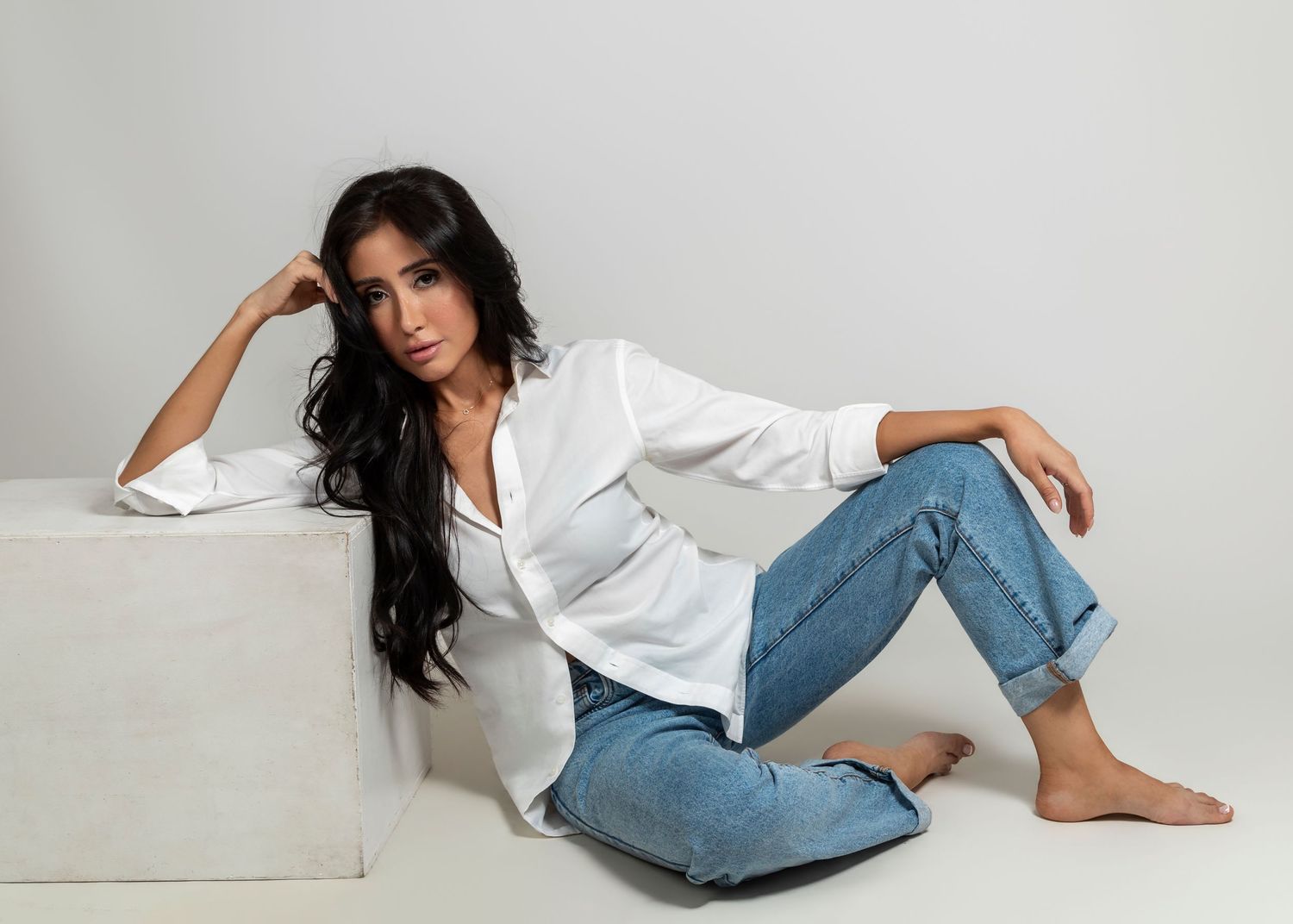 Woman in white blouse and blue jeans sitting on floor leaning against white cube in studio setting.