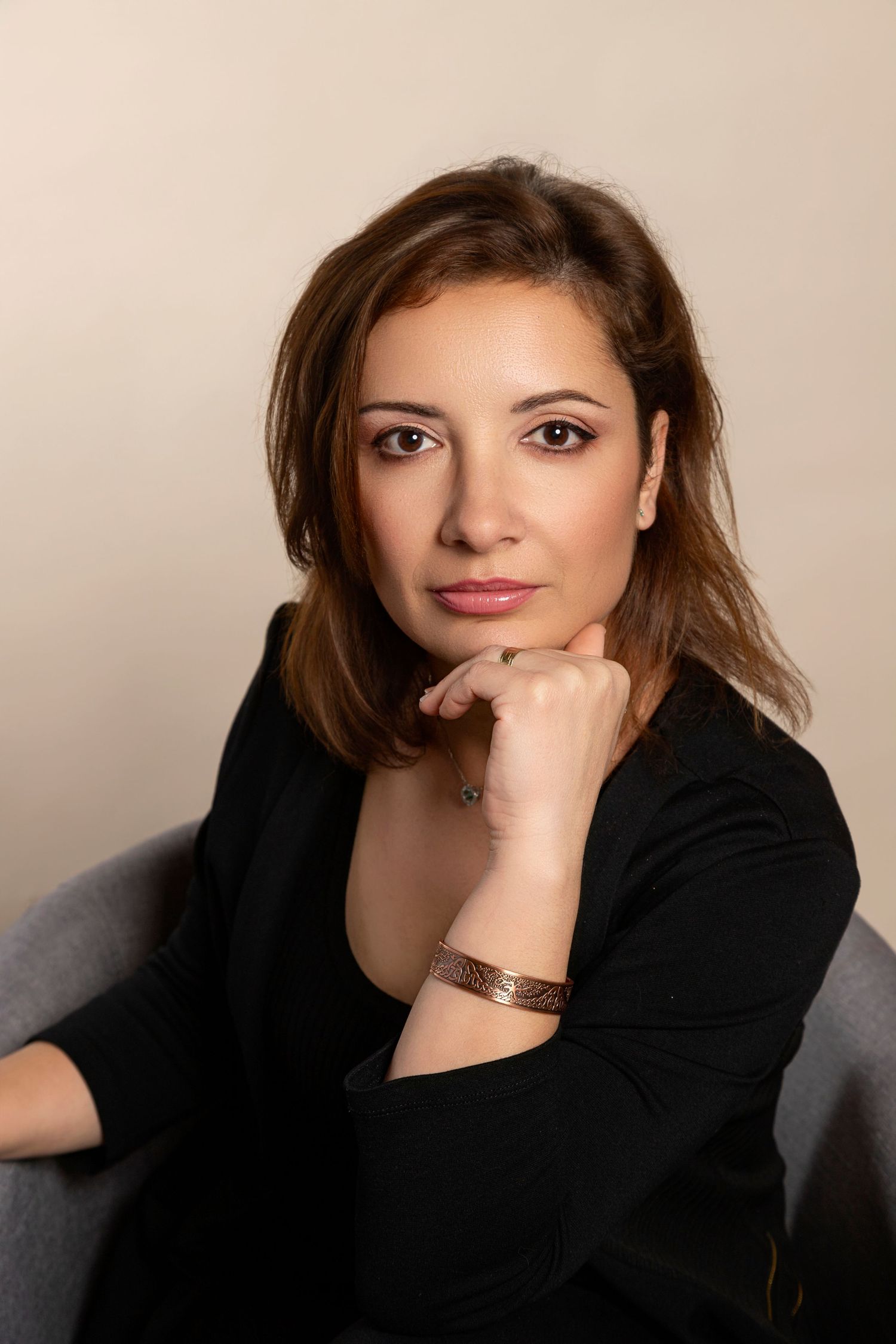 Professional portrait of a brunette woman in a black blazer sitting thoughtfully with hand on chin.