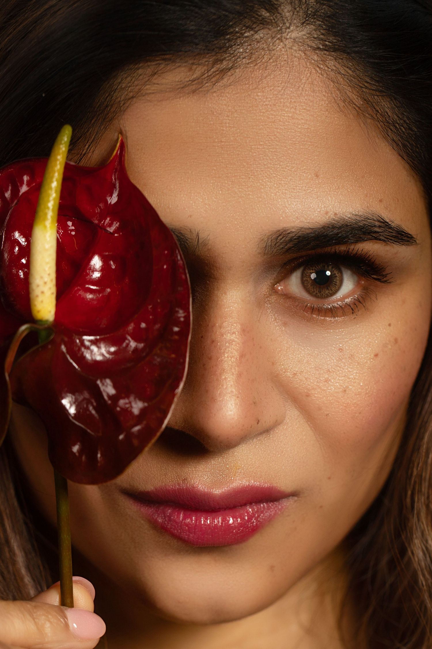 Close-up portrait of a woman holding a dark red anthurium flower over one eye, with natural makeup and brown eyes.