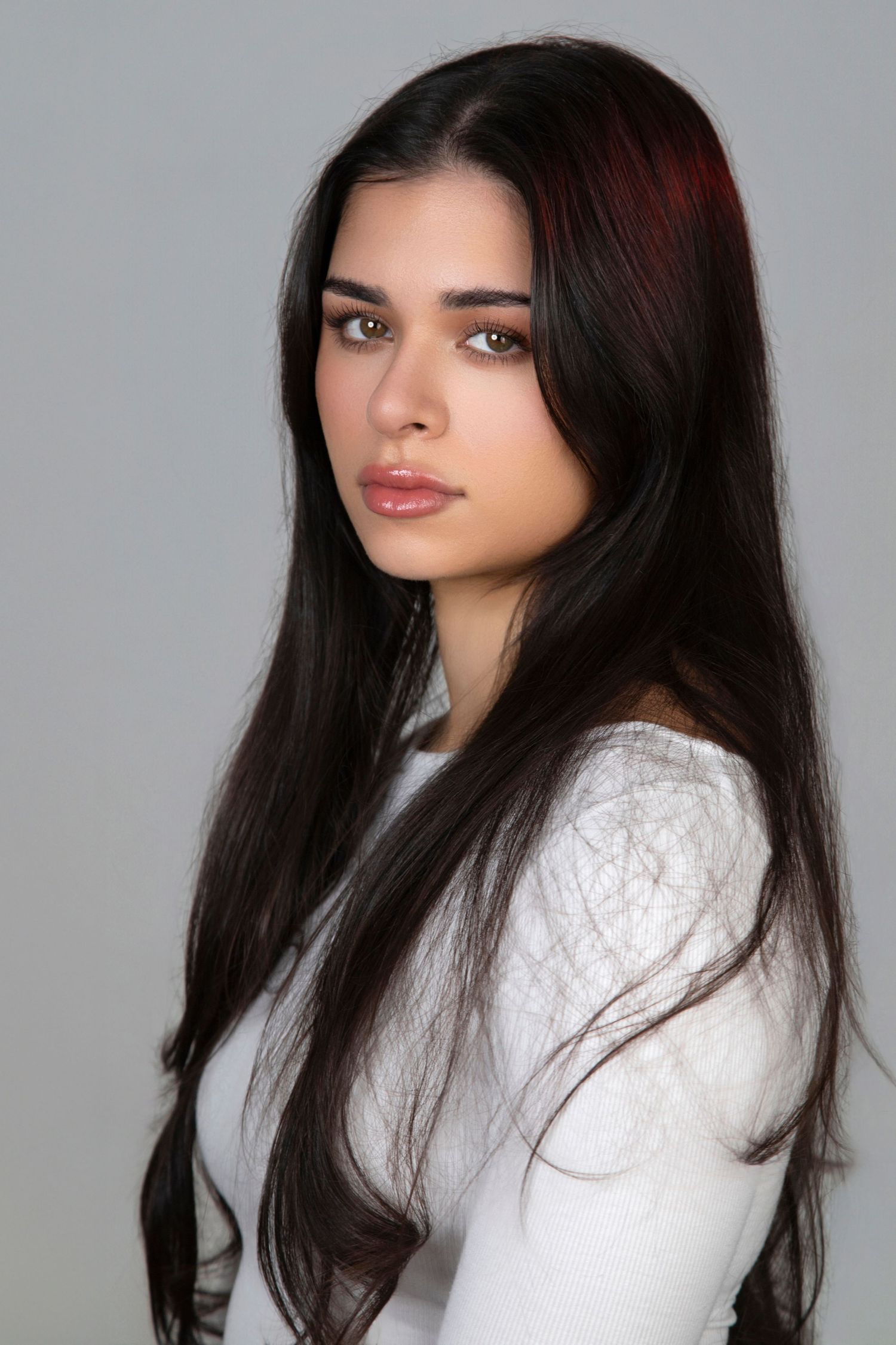 Young woman with long dark hair and green eyes wearing a white top poses against a gray background.