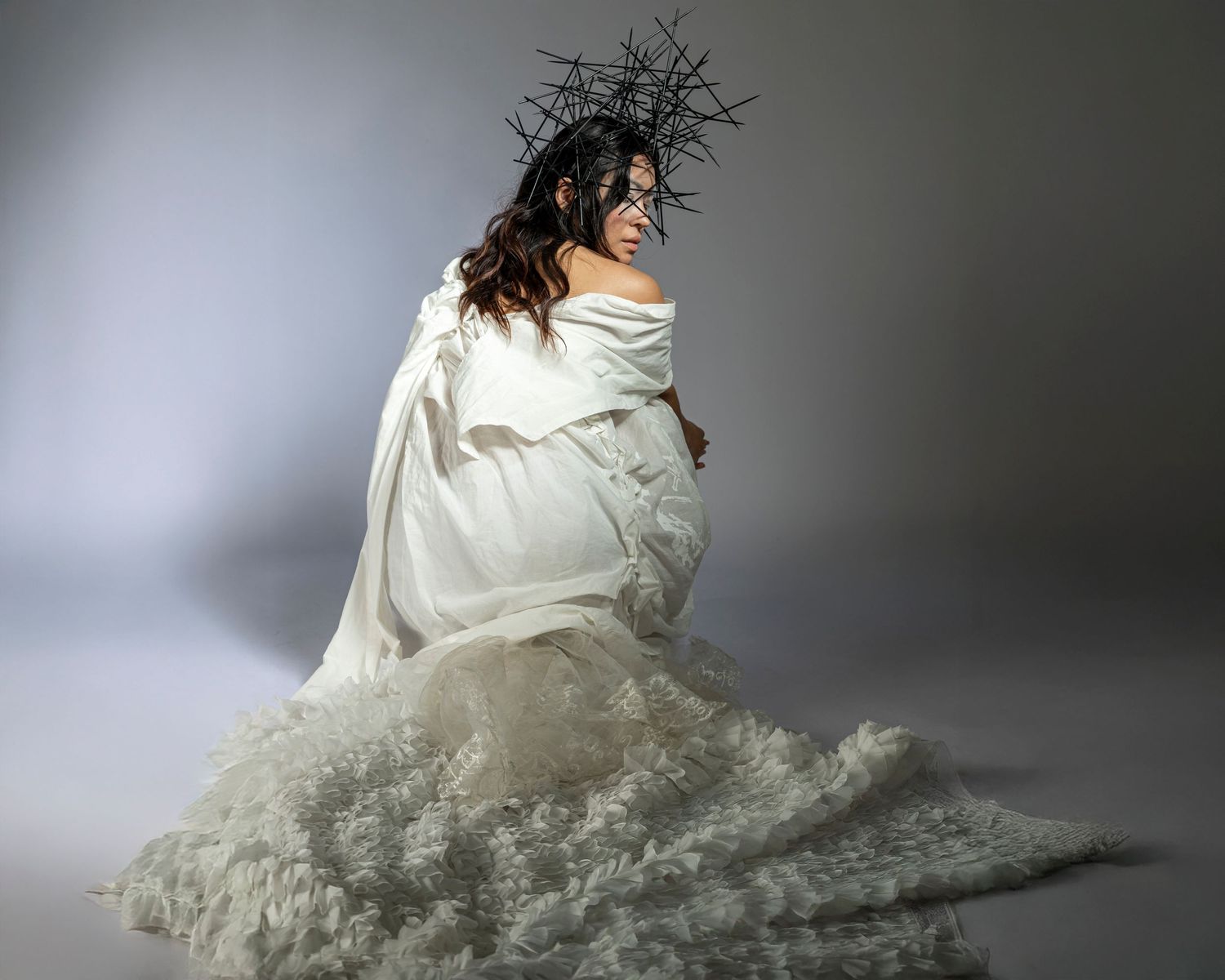 Woman in white flowing gown and dark thorn crown poses elegantly against a gray studio background.