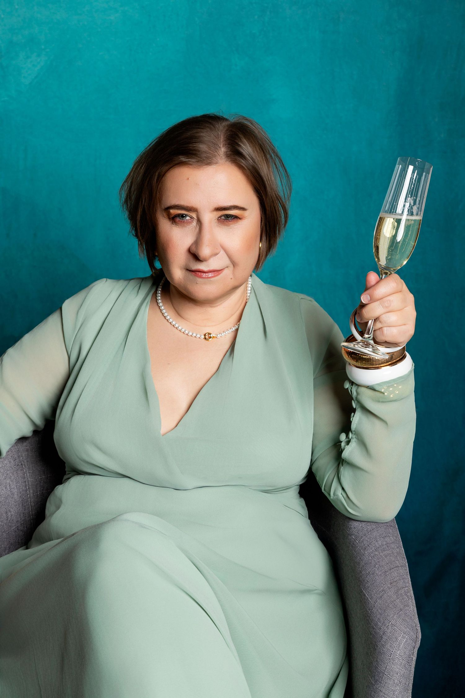 Woman in mint green dress holding champagne flute, seated in gray chair against teal background.