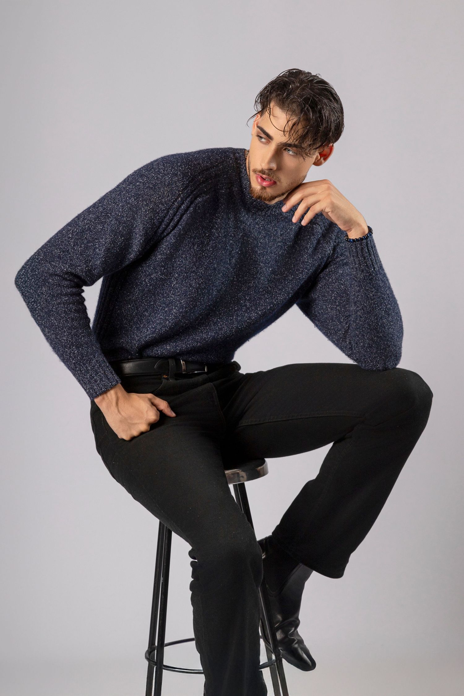 Man in blue knit sweater and black pants sitting on a stool in a studio setting.
