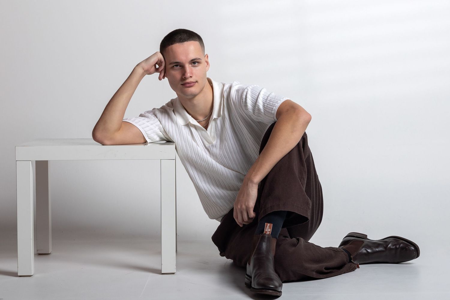 Young man in white polo and brown pants sitting on floor leaning against white table in studio setting.