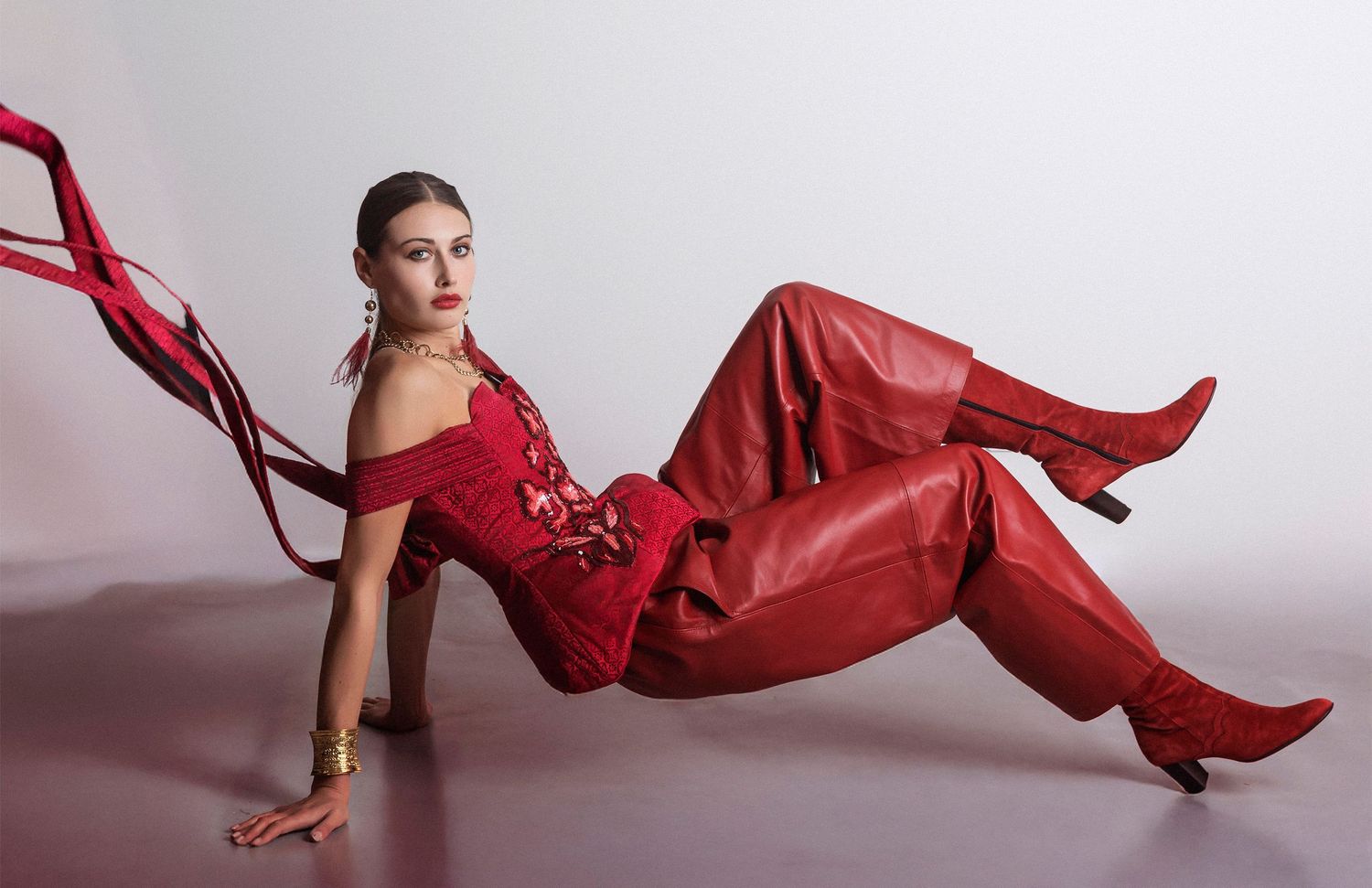 Woman in red leather outfit posing elegantly on floor with flowing red ribbon accessory and matching heeled boots.