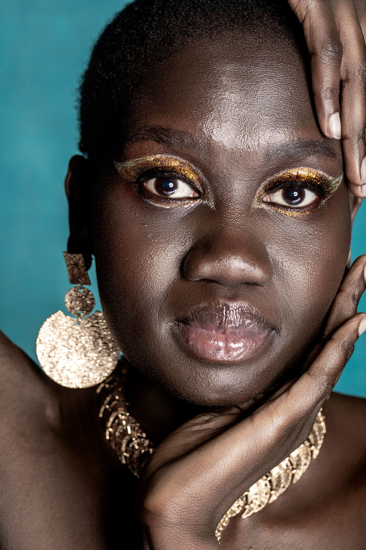 Close-up portrait of a woman with gold glitter eye makeup, large gold earrings, and bracelet on teal background.