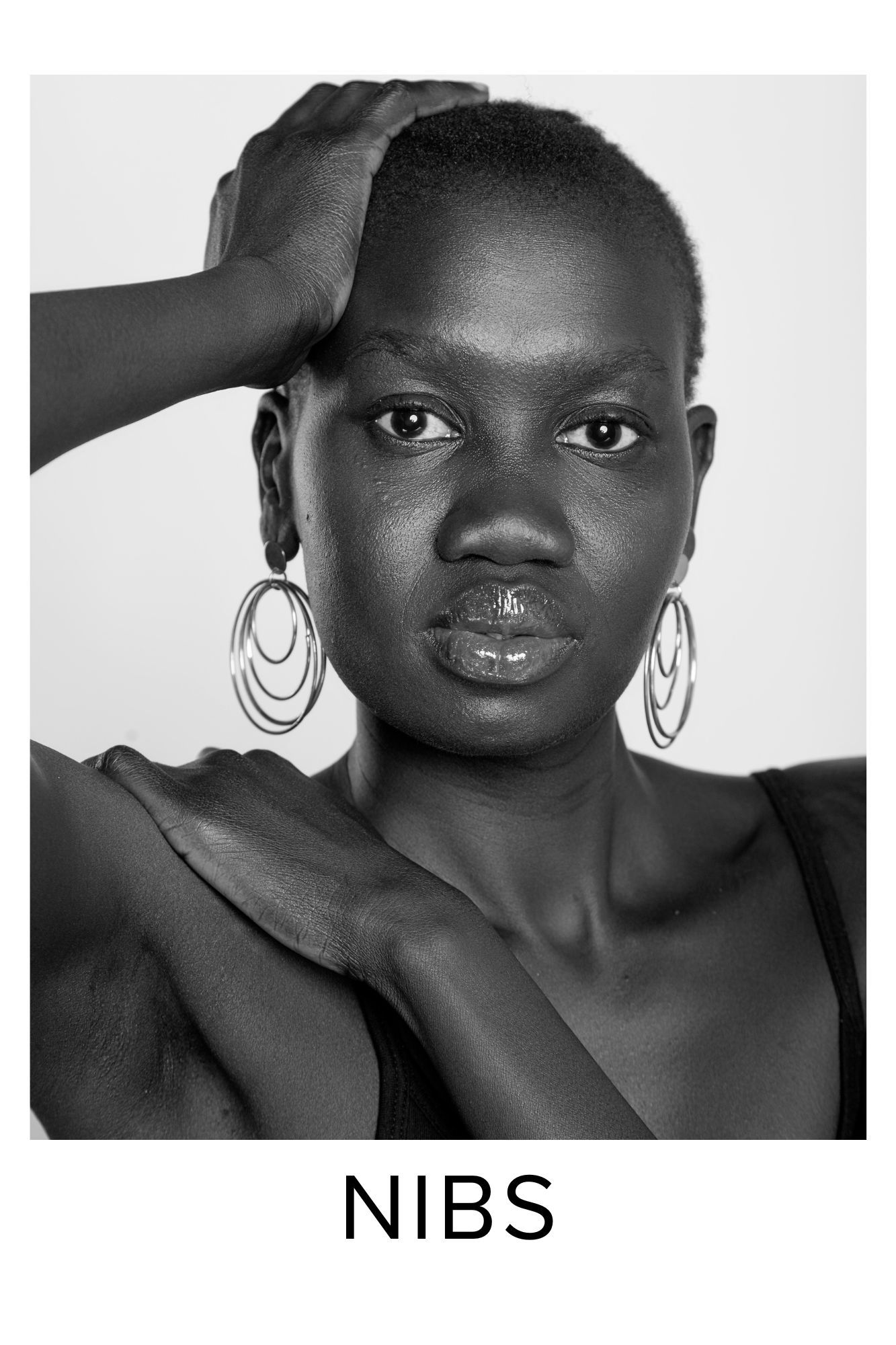 Black and white portrait of a model wearing large hoop earrings and a black top, with hand raised to head.