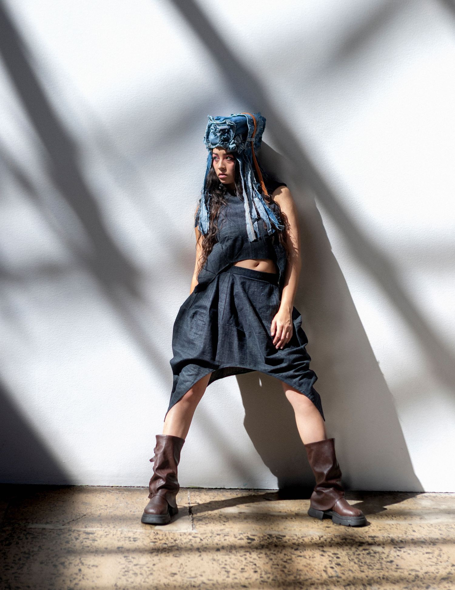Model in dark avant-garde outfit with blue headpiece, crop top, harem pants, and brown boots poses against white wall.