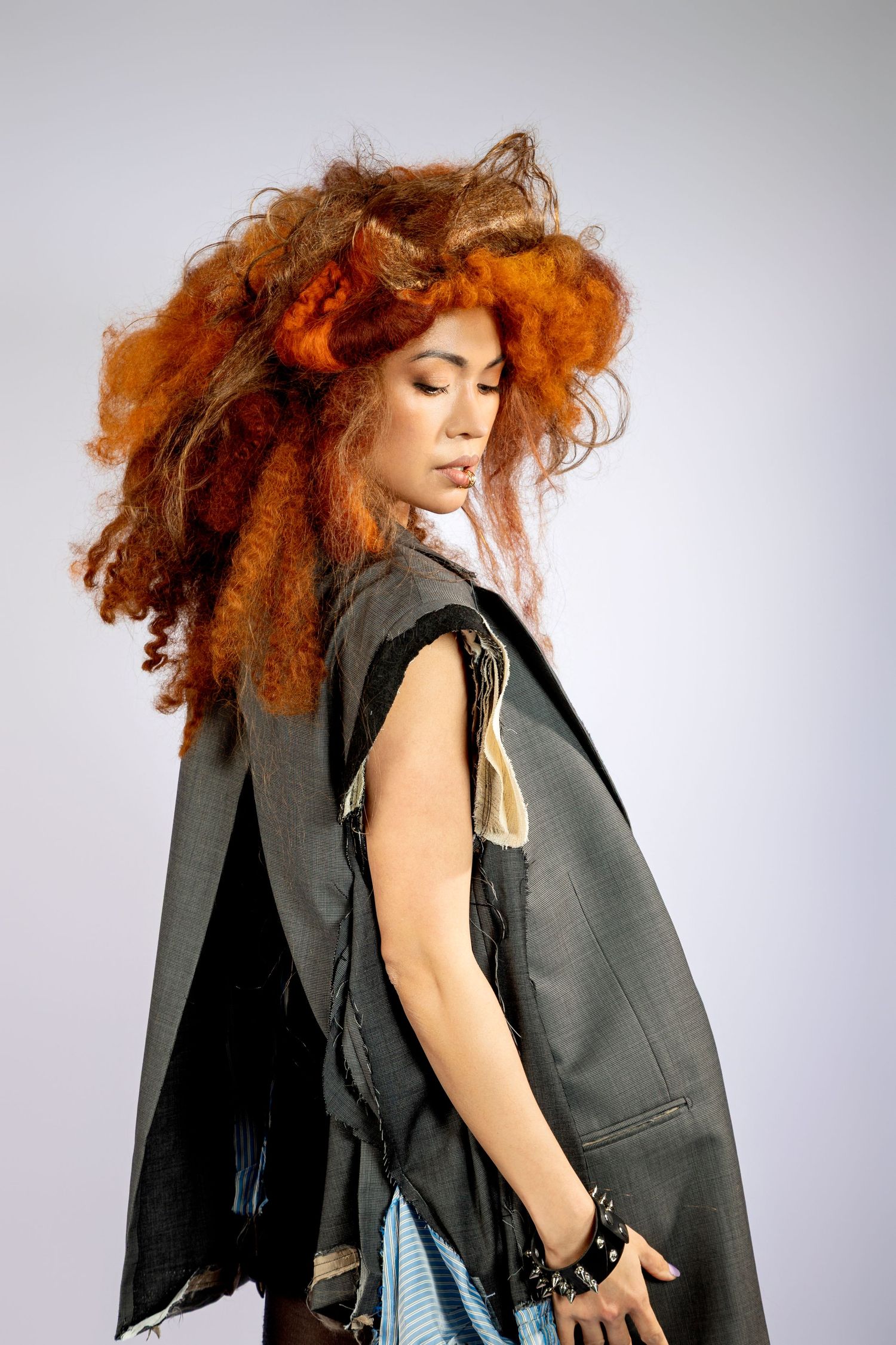 Woman with voluminous head piece wearing a dark cape-style outfit, posing dramatically against a gray background.