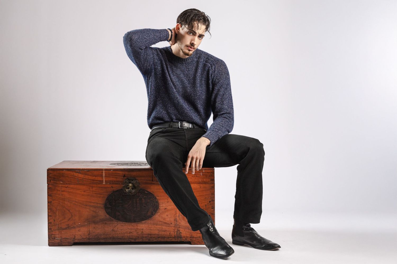 Man in blue sweater and black pants sitting on wooden chest with hand behind head against white background.