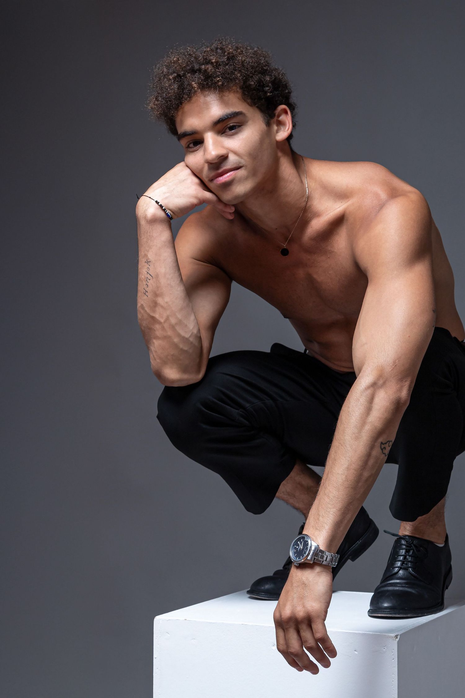 Shirtless muscular young man with curly hair sitting on white cube, wearing black pants and watch, against gray background.
