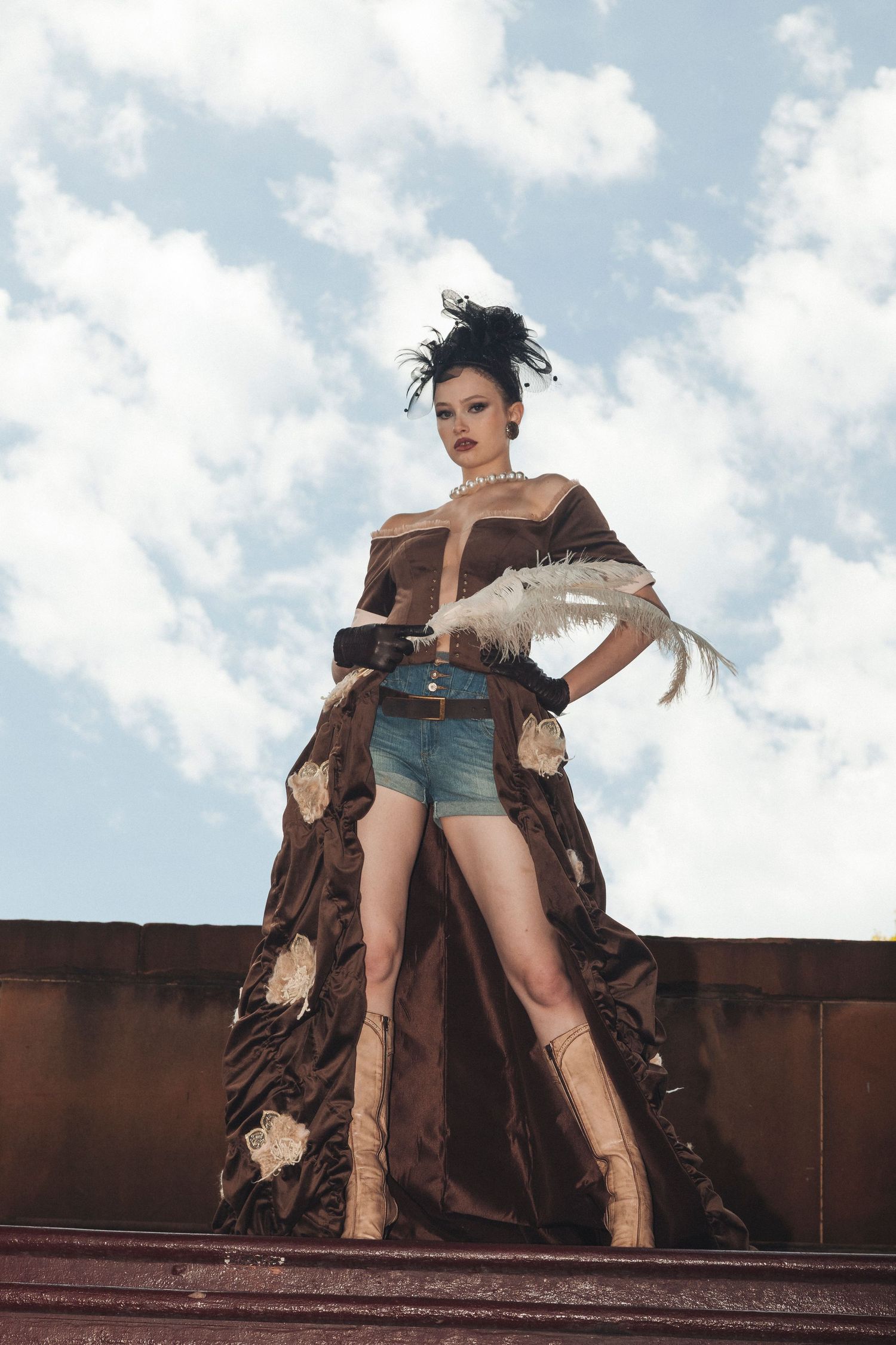 Woman in brown floral gown with denim shorts and feather accessory posing outdoors against cloudy sky.