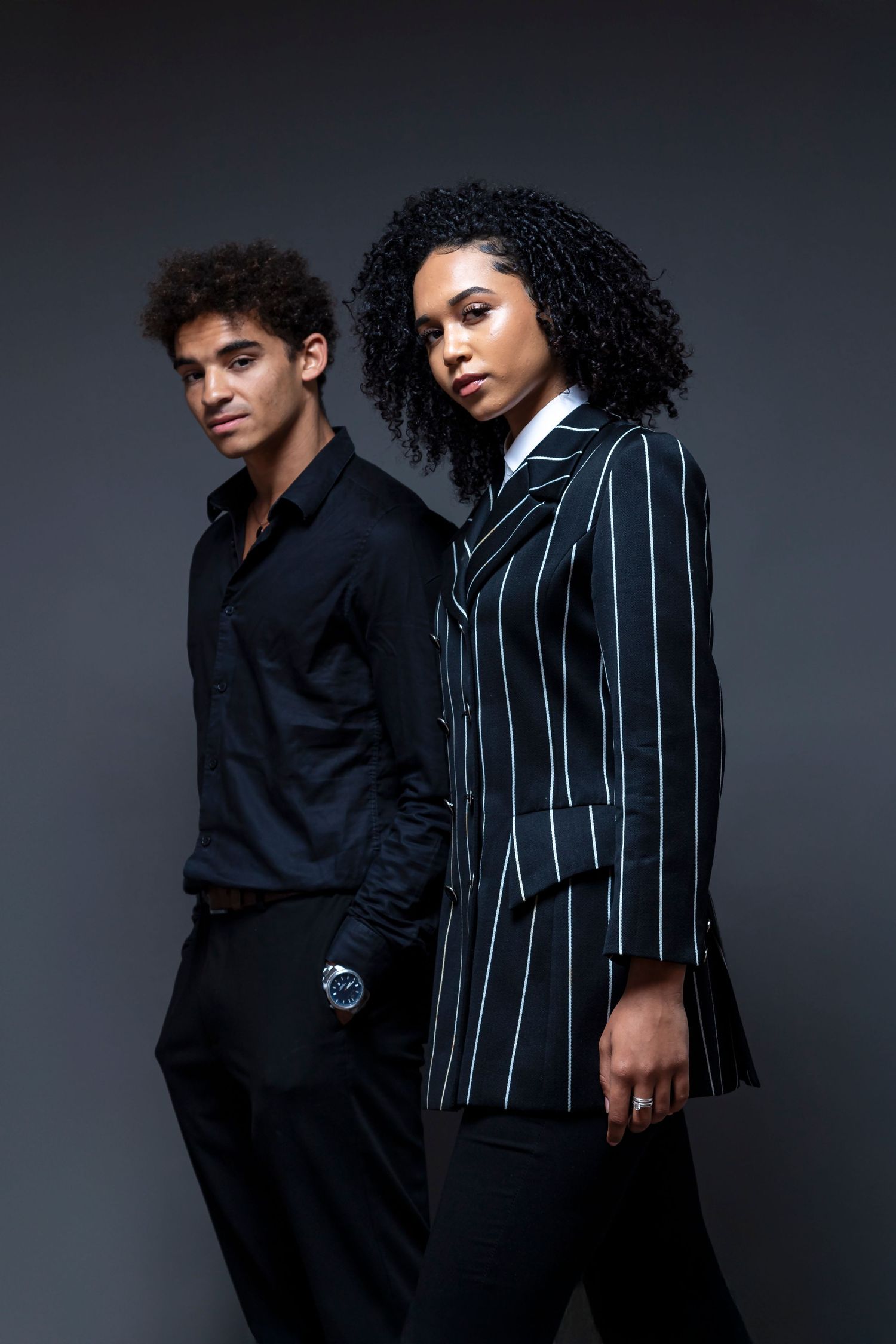 Two stylish models pose against dark background, woman in pinstripe blazer, man in black shirt with watch.