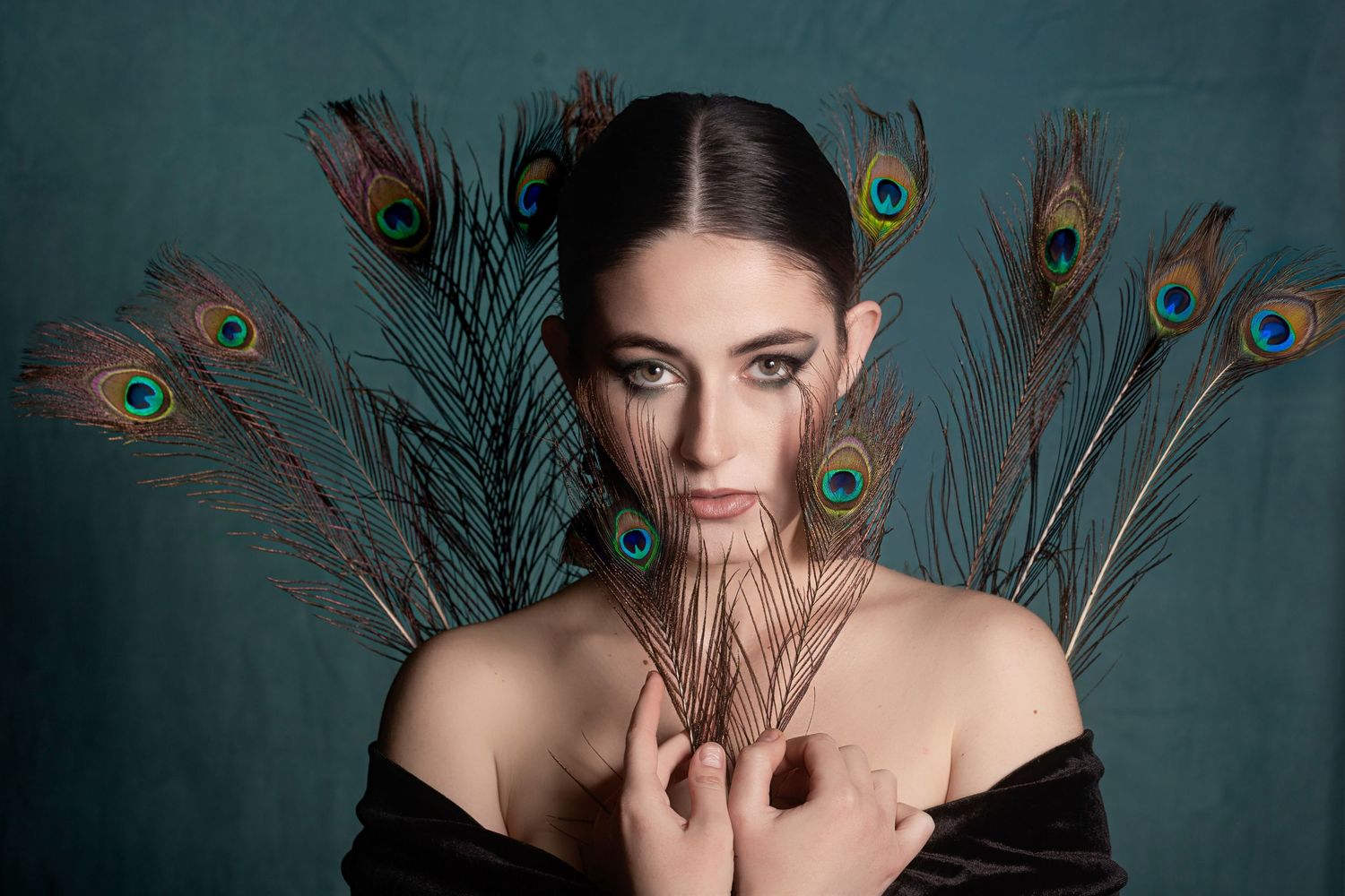 Young woman with dark hair holds peacock feathers in a dramatic portrait against a teal background.