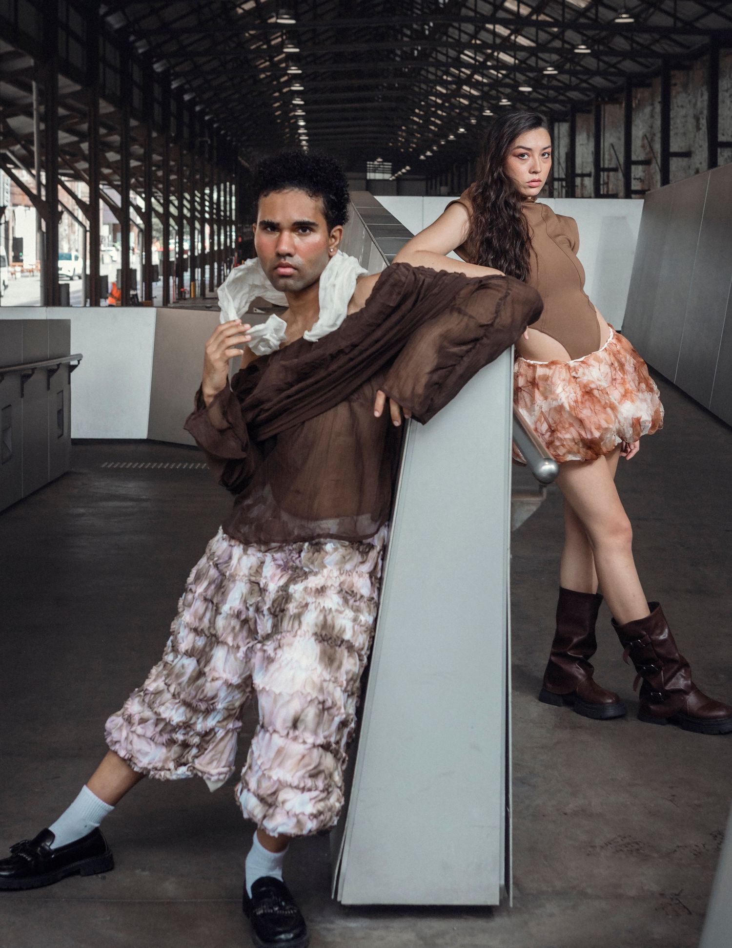 Two models pose in an industrial warehouse wearing brown sheer avant-garde tops and tie-dye shorts with loafers and boots.