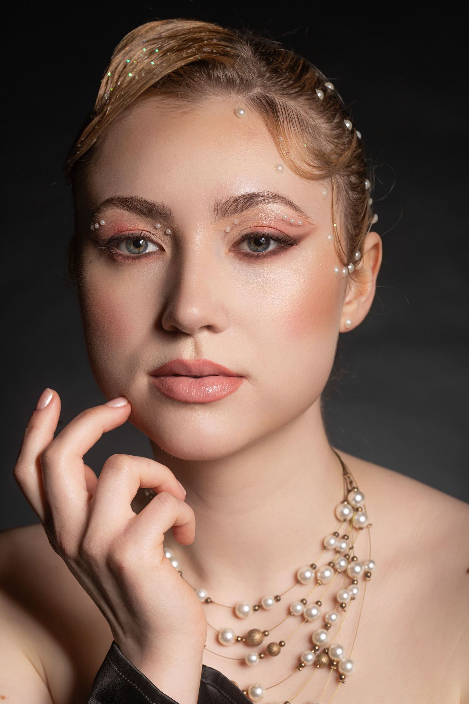 Young woman with pearl hair accessories and necklace, elegant makeup, posing gracefully against dark background.