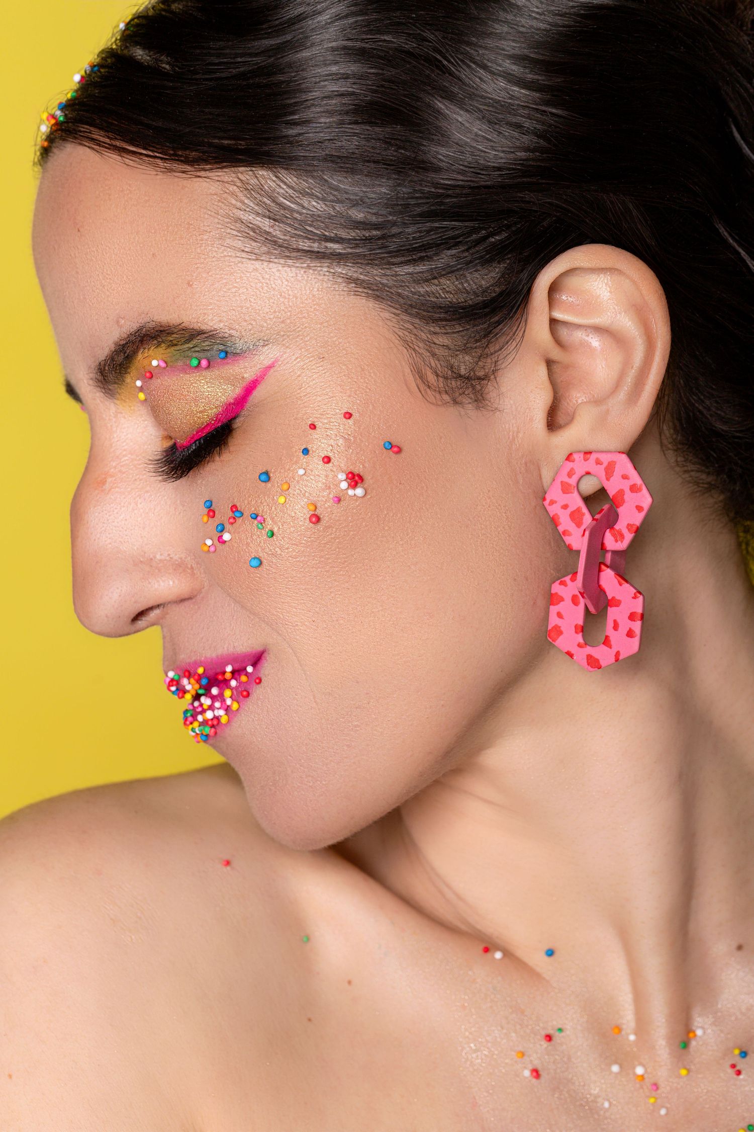 Woman with colorful glitter makeup, pink donut earrings, and sprinkles on face against yellow background.