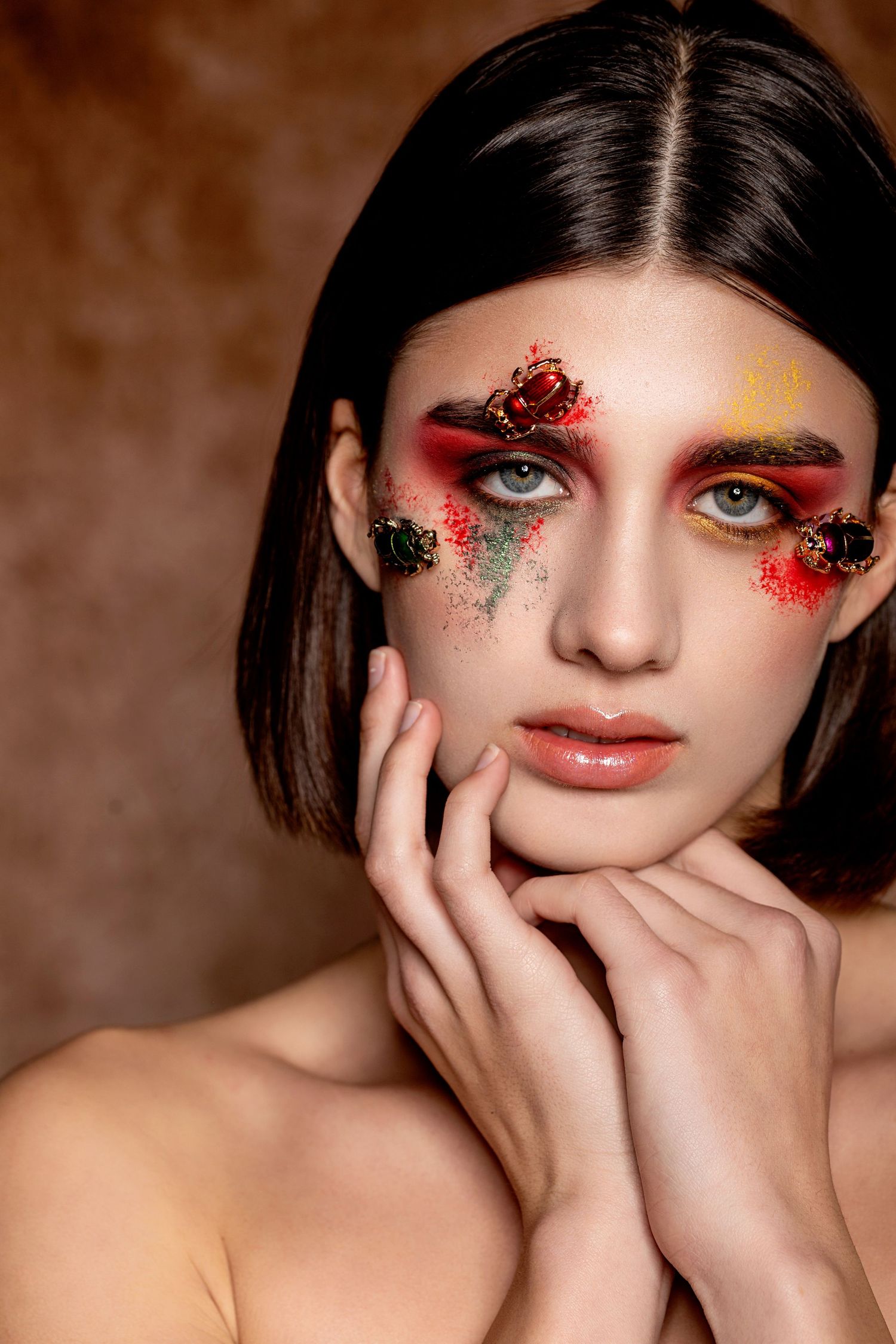 Woman with avant-garde makeup featuring red eyeshadow, colorful gems, and artistic face paint poses gracefully.