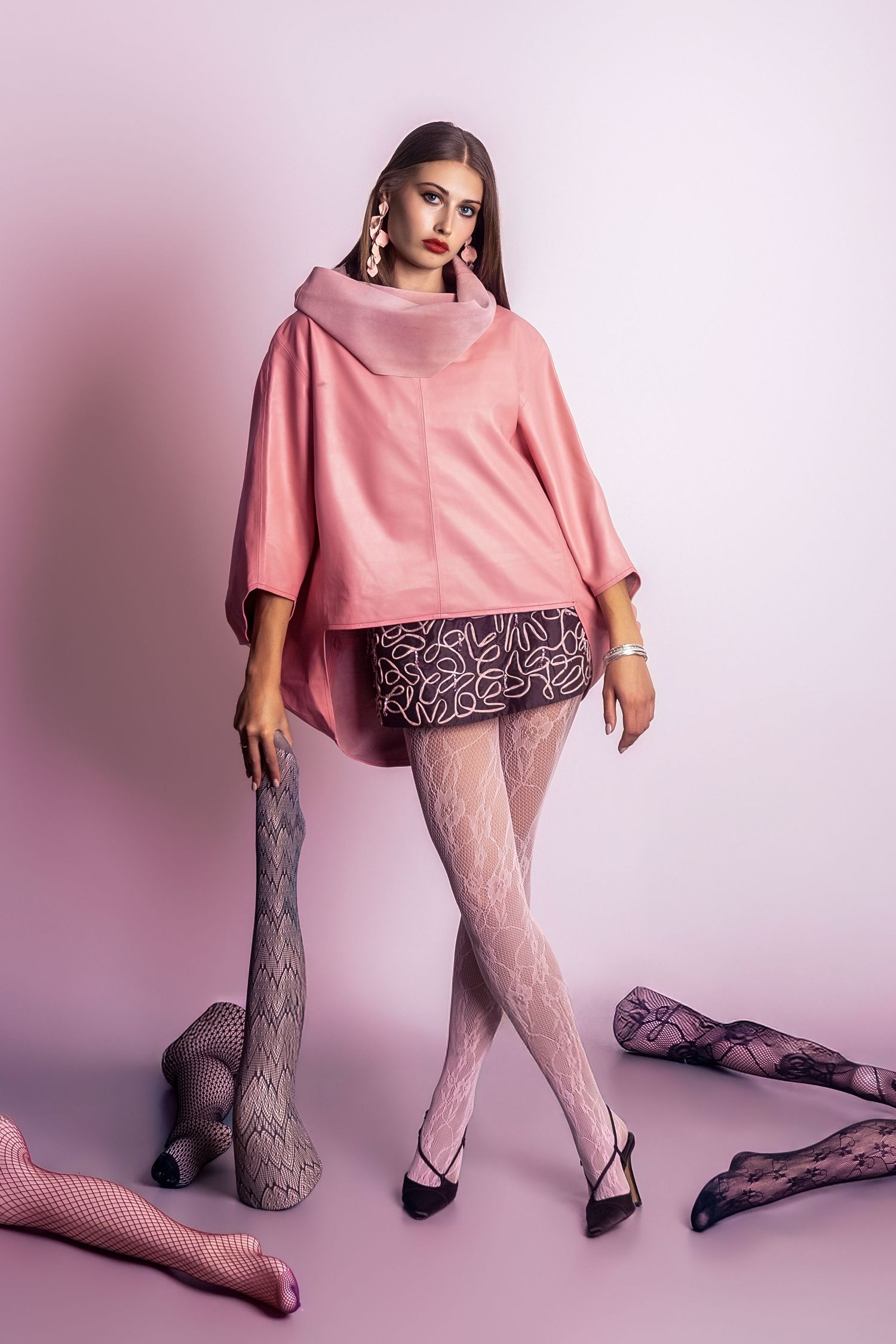Woman in pink oversized top, mini skirt, and white patterned tights poses among scattered hosiery on pink background.