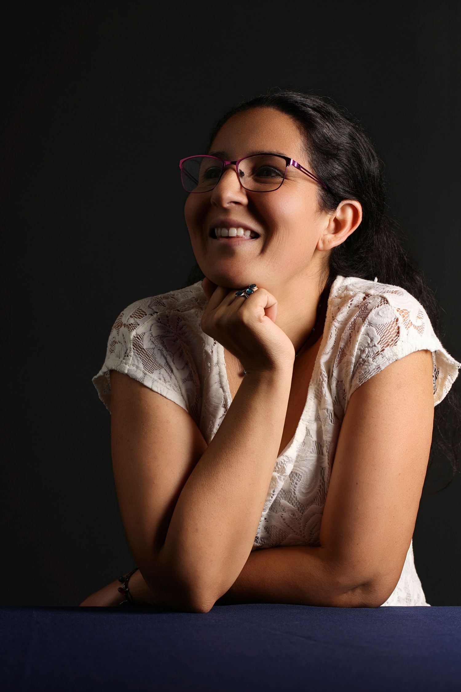 Smiling young woman with glasses wearing a white lace top, posing against a dark background.