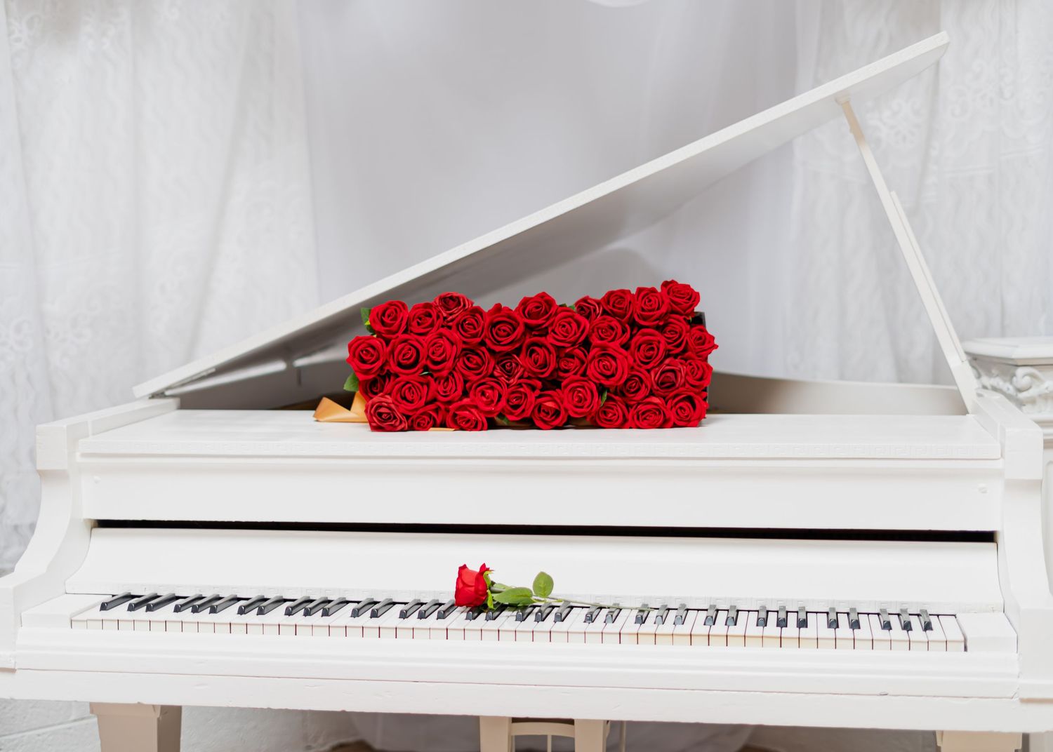 Bouquet of red roses arranged on a white grand piano with sheer white curtains behind.
