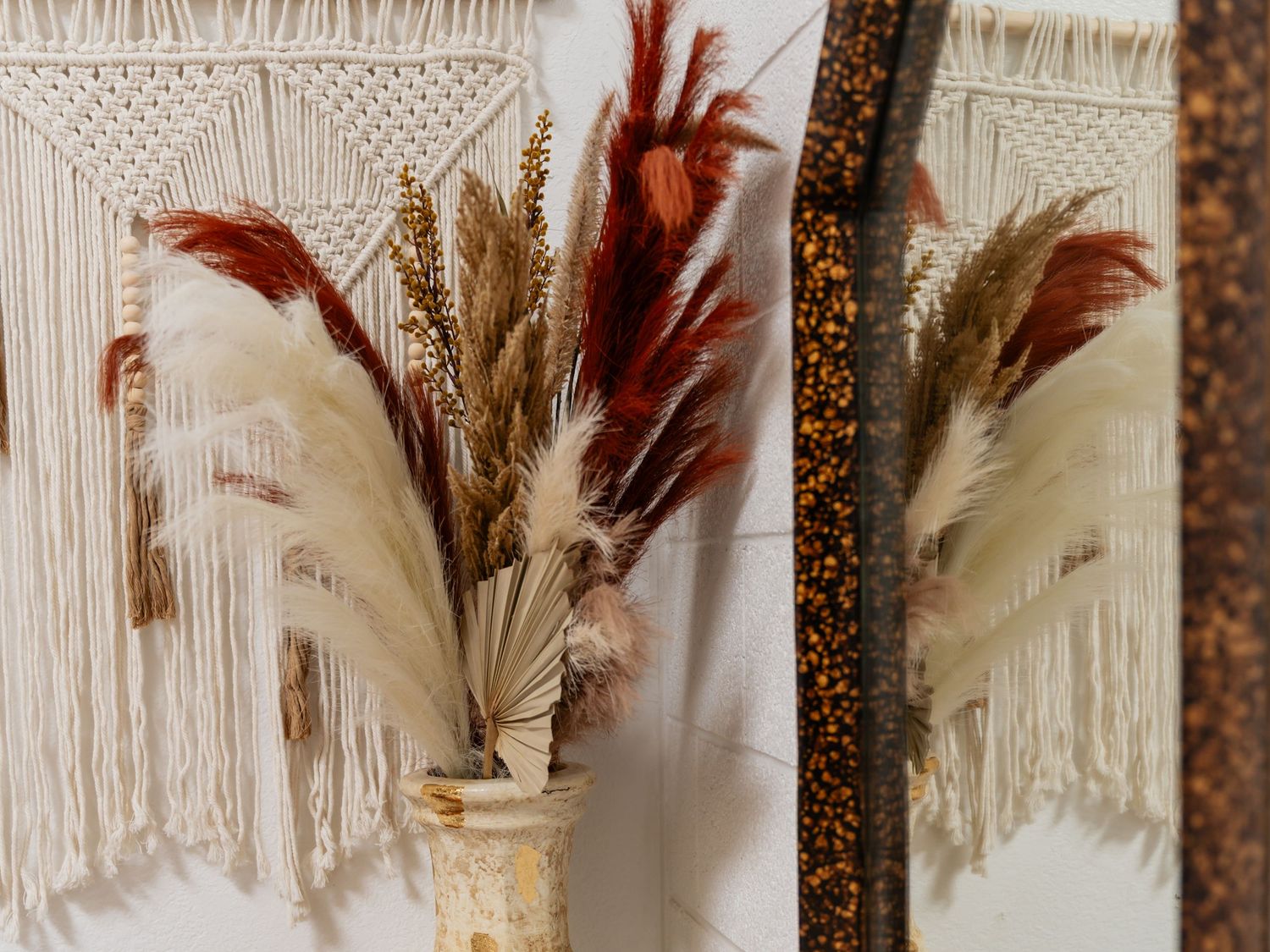 Bohemian macrame wall hanging with dried pampas grass and wheat stems arranged in a tall vase beside a mirror.
