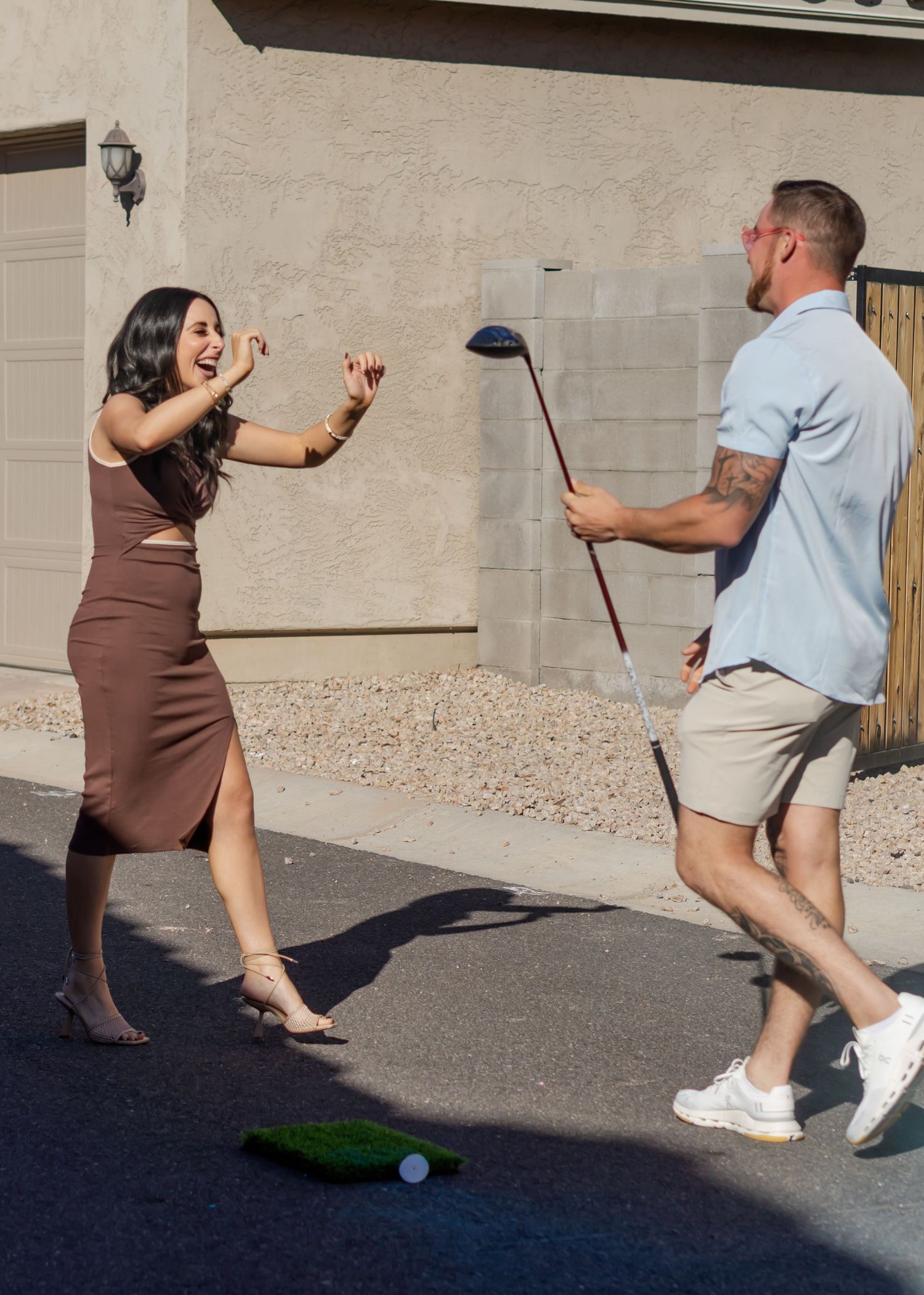 Couple laughing and playing golf together in a suburban driveway on a sunny day.