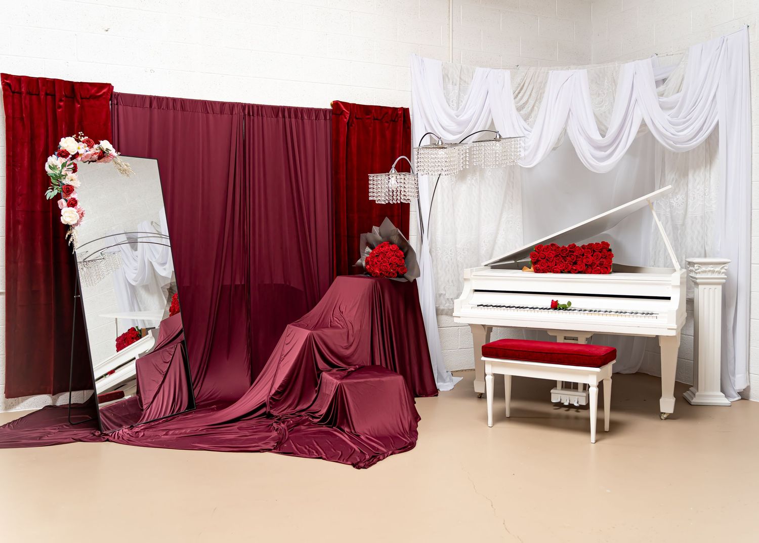Elegant photo studio setup with white grand piano, red roses, burgundy drapes, and a decorative mirror on beige flooring.
