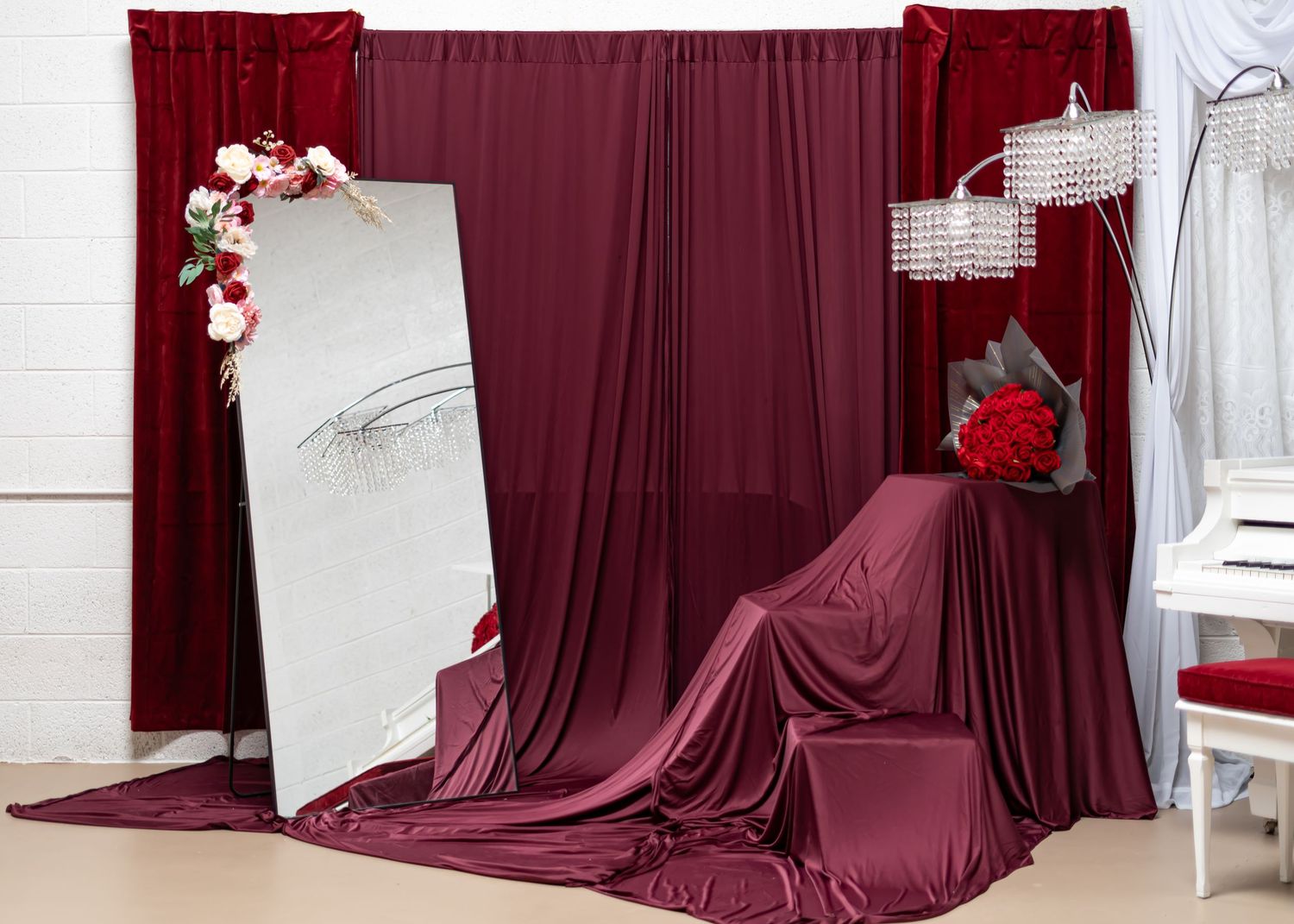 Elegant burgundy drape backdrop setup with chandelier, mirror, and floral accents for photography.
