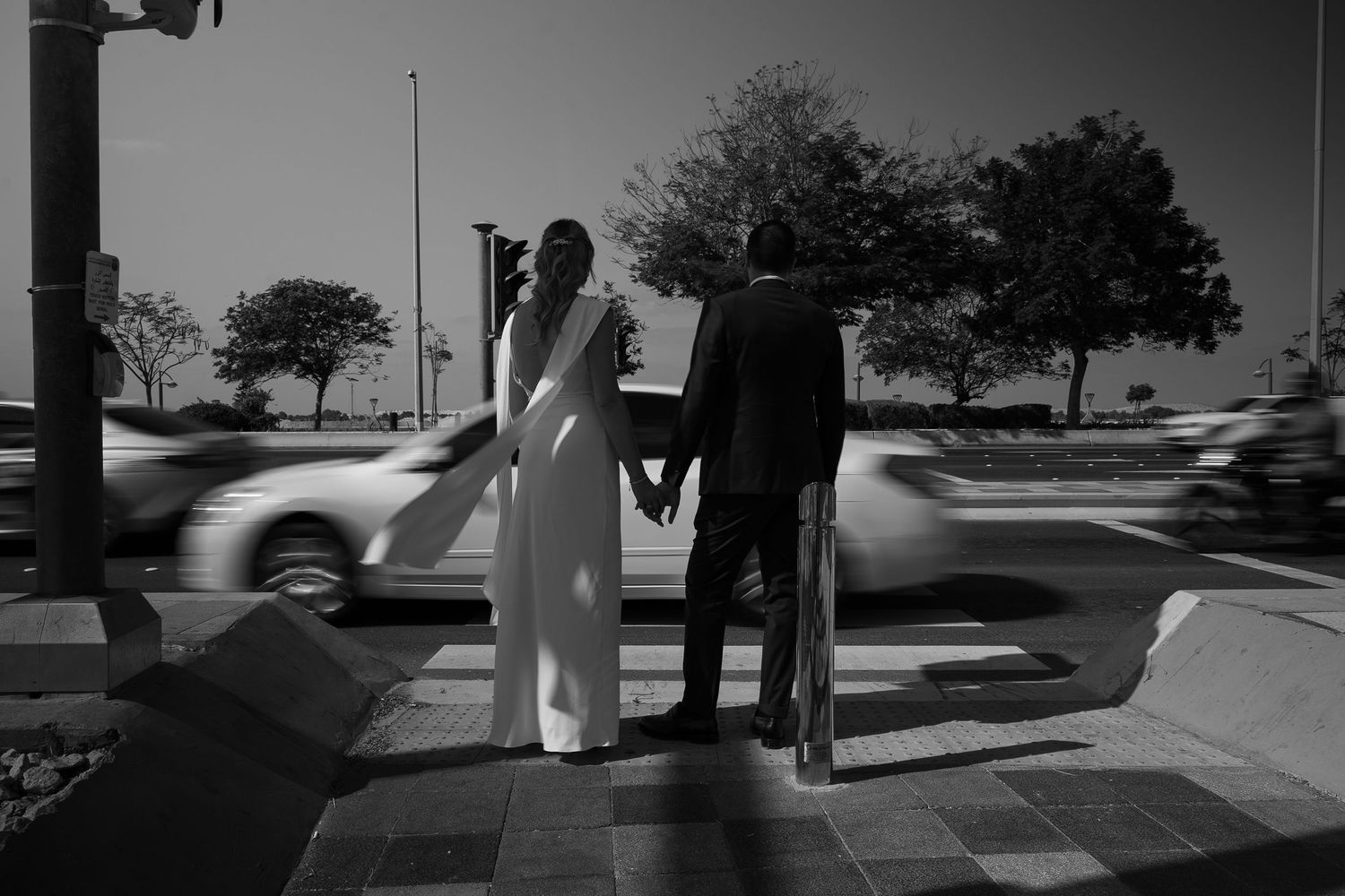 Black and white photo of a bride and groom holding hands at a crosswalk with blurred traffic passing by.