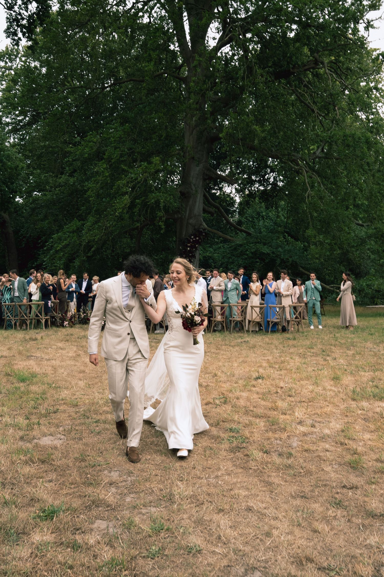 Bride and groom walk joyfully through outdoor wedding ceremony aisle surrounded by guests under large green trees.