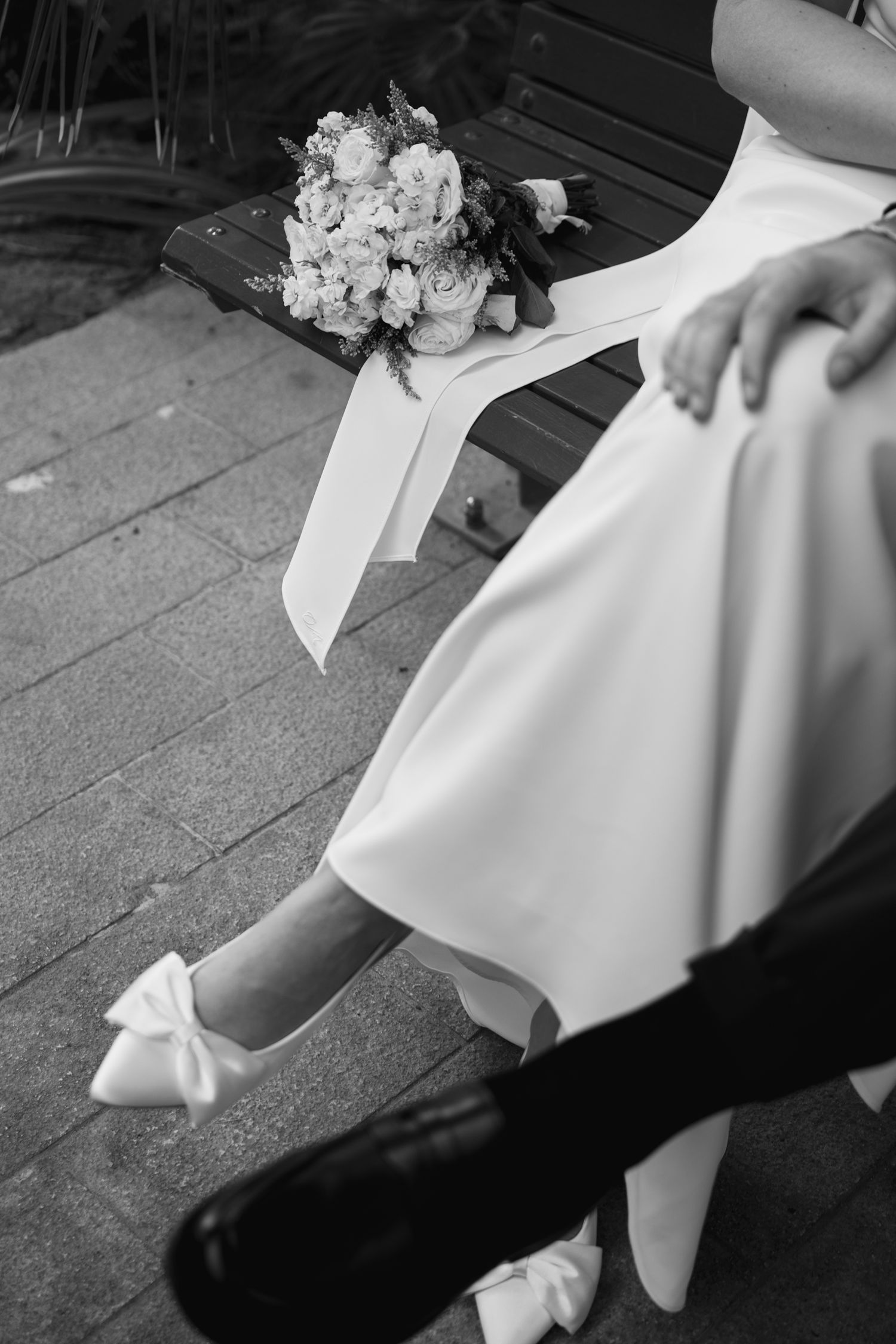 Black and white photo of a bride in white dress and bow heels sitting on bench with wedding bouquet.