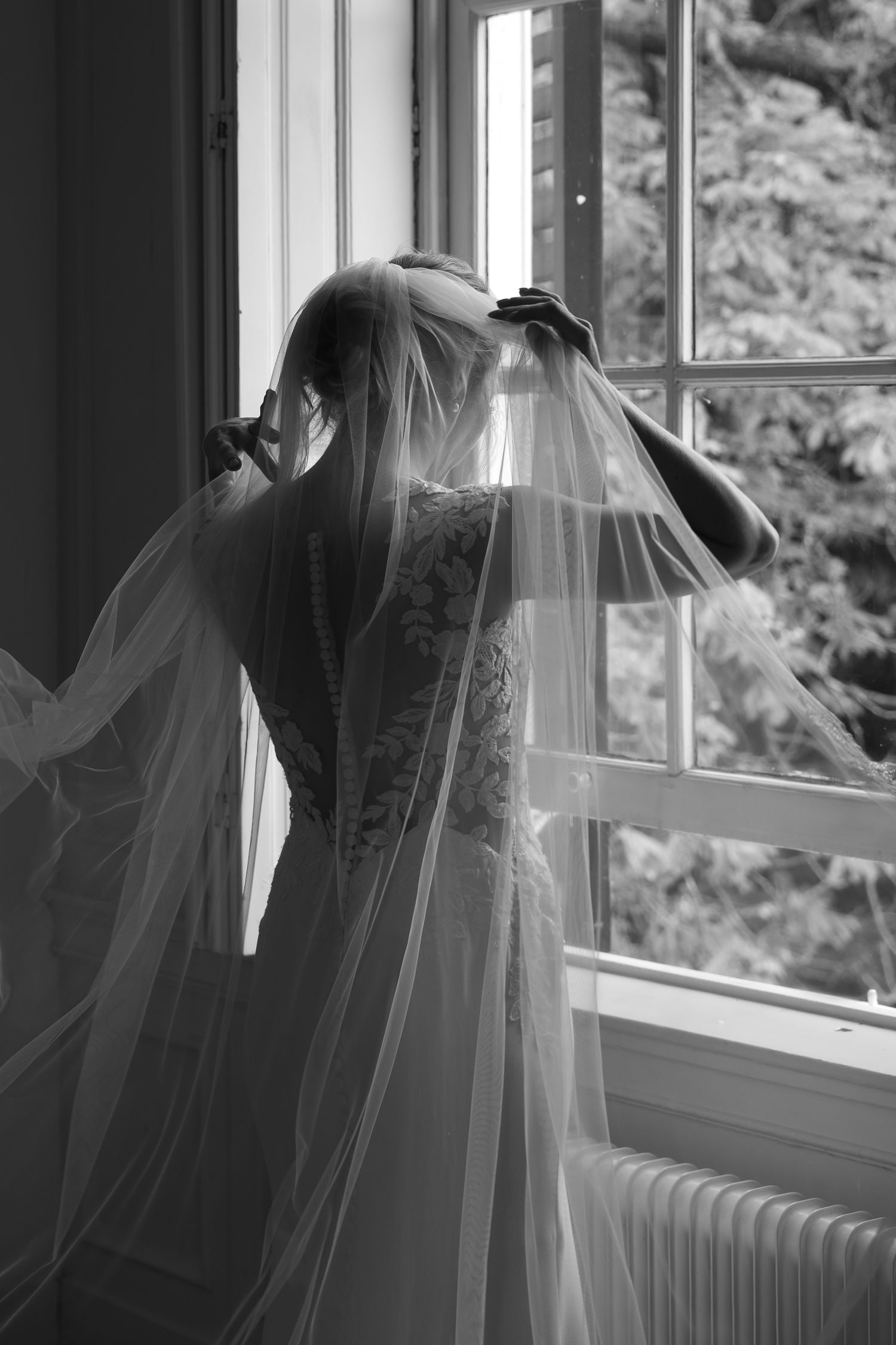 Black and white photo of a bride in lace gown adjusting her veil by a window with trees visible outside.