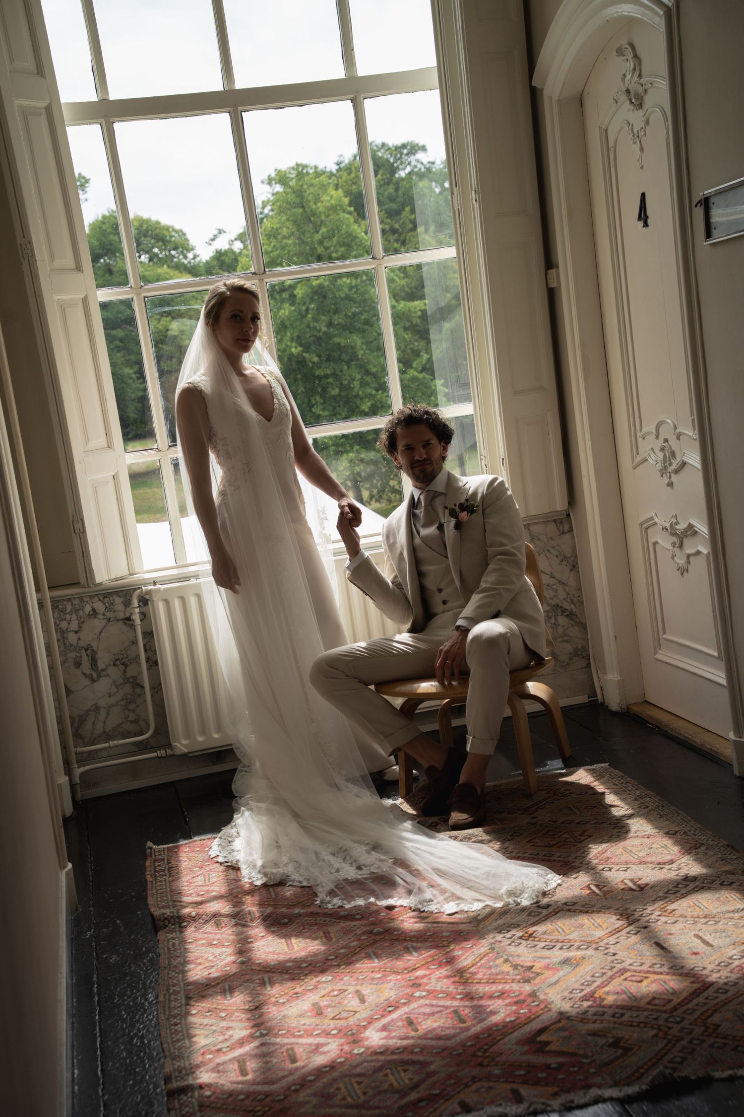 Bride in white gown and groom in beige suit hold hands by a sunlit window in an elegant venue.